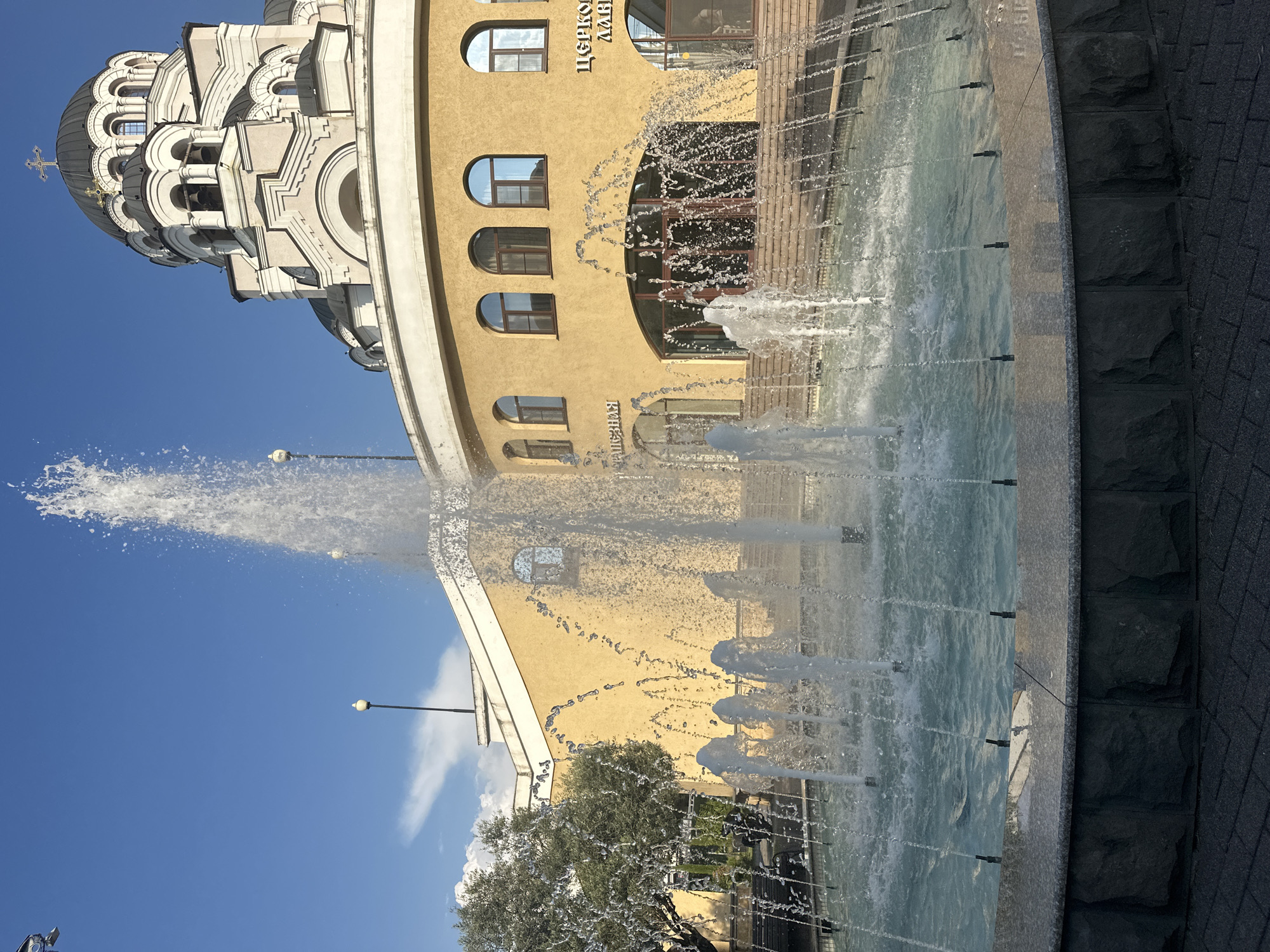The photograph captures a sunny day in Sochi, Russia, featuring a modern church complex with a prominent fountain in the foreground. The church, characterized by its domes and bell towers, is constructed with a light-colored stone and a dark roof. A portion of the yellow facade of an adjacent building is visible, adorned with arched windows. The fountain, a central element, is actively spraying water, with multiple jets creating a dynamic visual effect against the clear blue sky. The surrounding area includes paved walkways and greenery, suggesting a public square or park setting. No people are visible in the image.