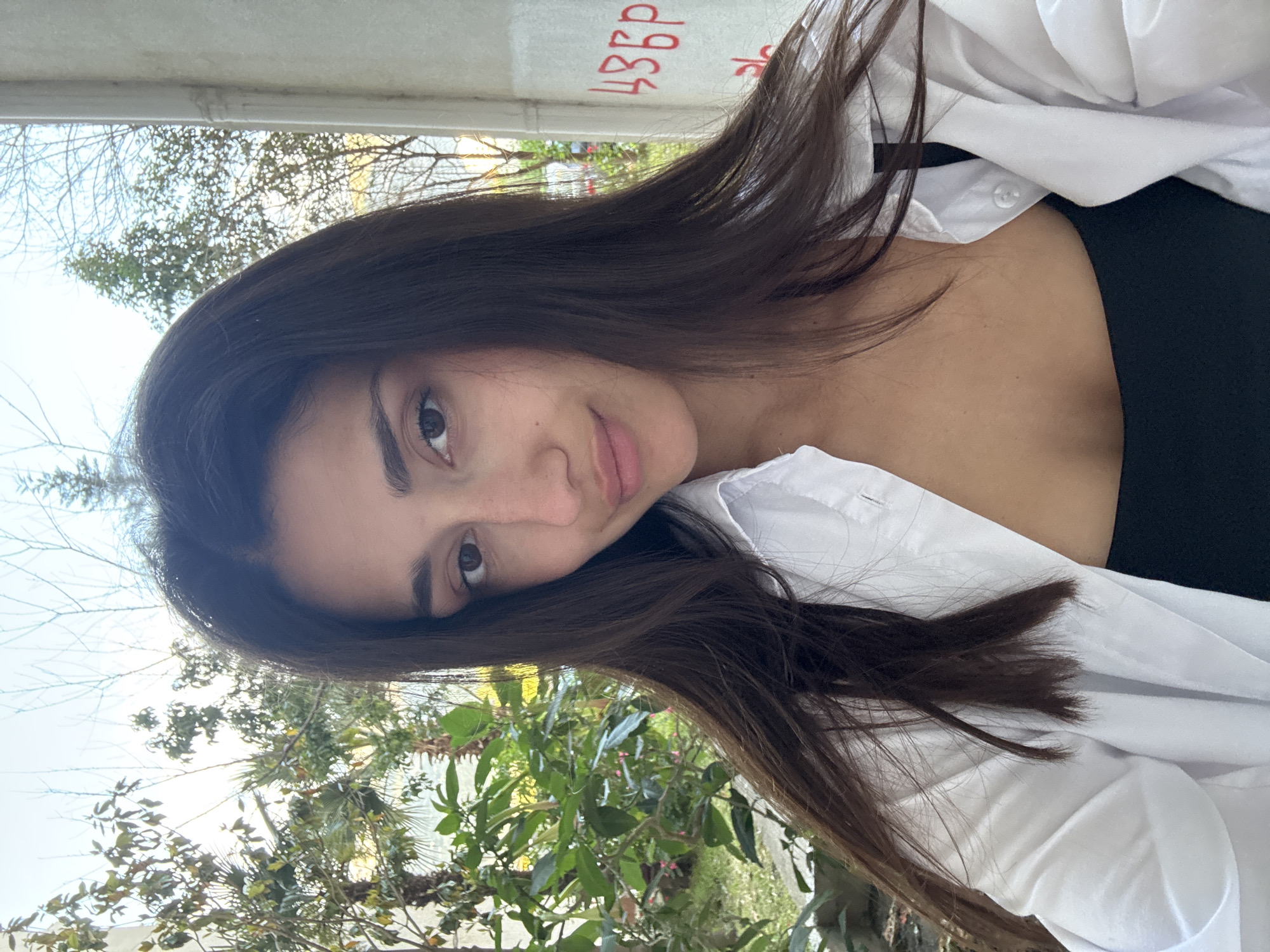 A woman with long, dark brown hair is posing for a selfie. She is wearing a black top underneath a partially unbuttoned white shirt. The background shows greenery, possibly a tree or bush. In the upper part of the frame there is some graffiti and branches. The selfie appears to have been taken outdoors, possibly in Bar, Montenegro, based on the location.