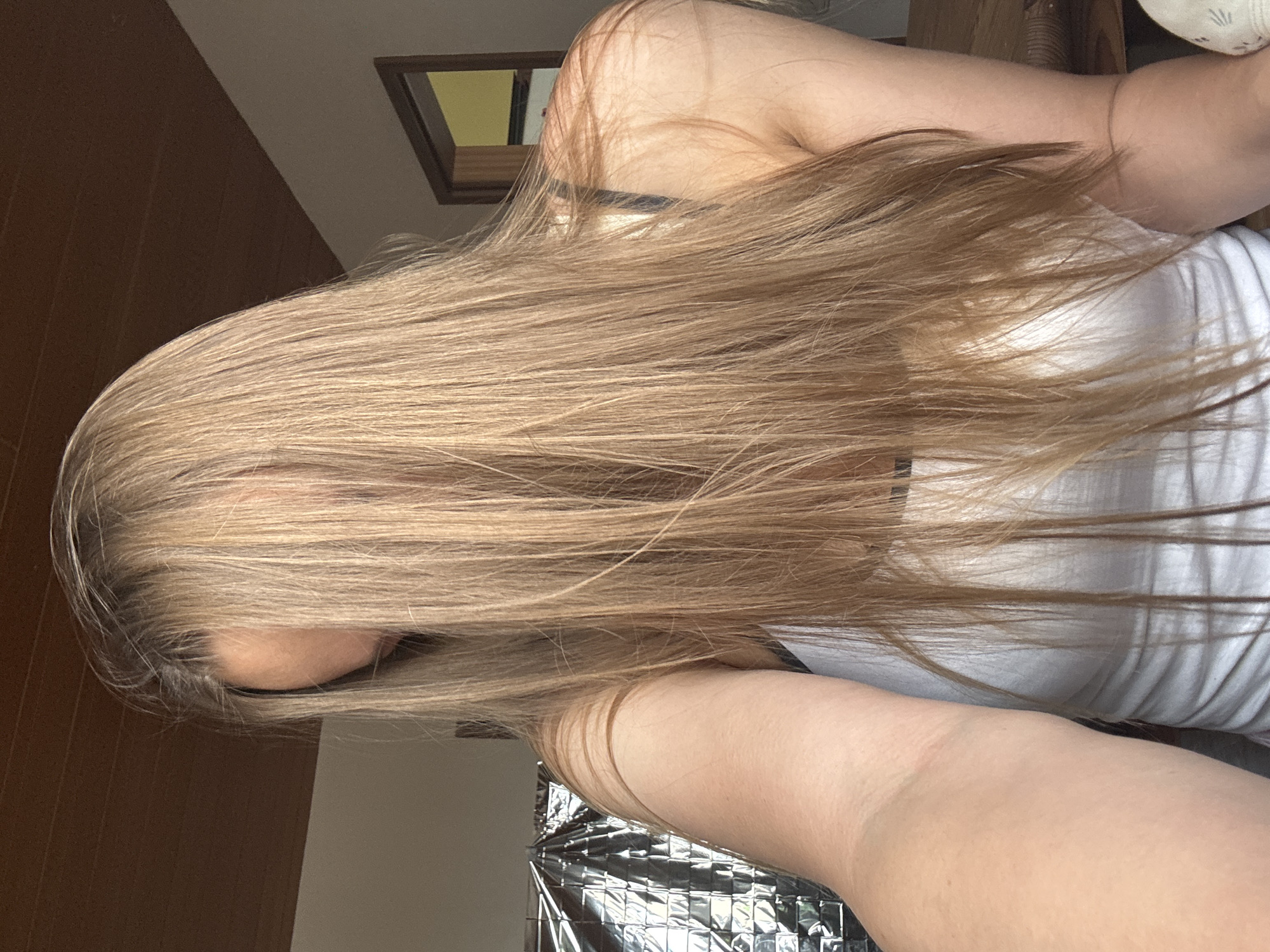 The image shows a person with long, blonde hair in a close-up shot. The person is wearing a white tank top. The background is blurred but suggests an indoor setting with wooden elements on the ceiling and a door frame visible. The lighting appears to be natural. There is no identifiable location in the city of Überherrn, Germany.