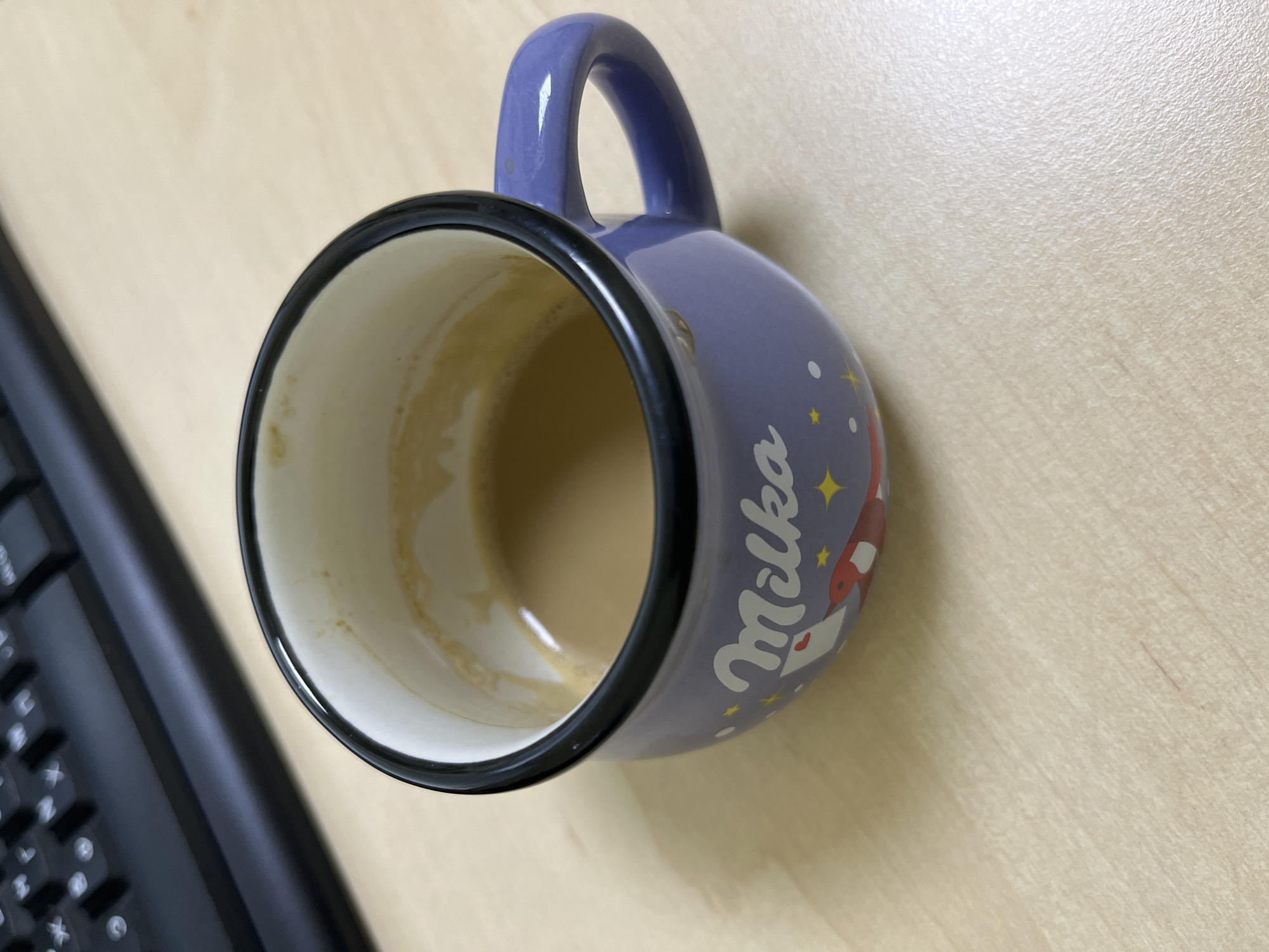A purple mug with the word "Milka" on it, containing a light brown liquid, sits on a light brown wooden surface. A portion of a black keyboard is visible in the upper left corner of the image.