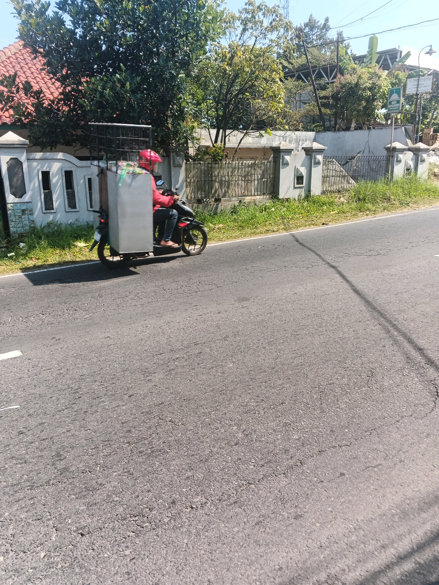 A person wearing a red jacket and red helmet rides a motorcycle with a large grey cargo box attached to the back. The motorcycle is traveling on a paved road with a white line marking the edge of the lane. The background shows a residential area with a white fence, green grass, and trees. The sky is clear and blue. The scene appears to be in Bandungan, Indonesia, as indicated by the presence of typical Indonesian residential architecture and vegetation.