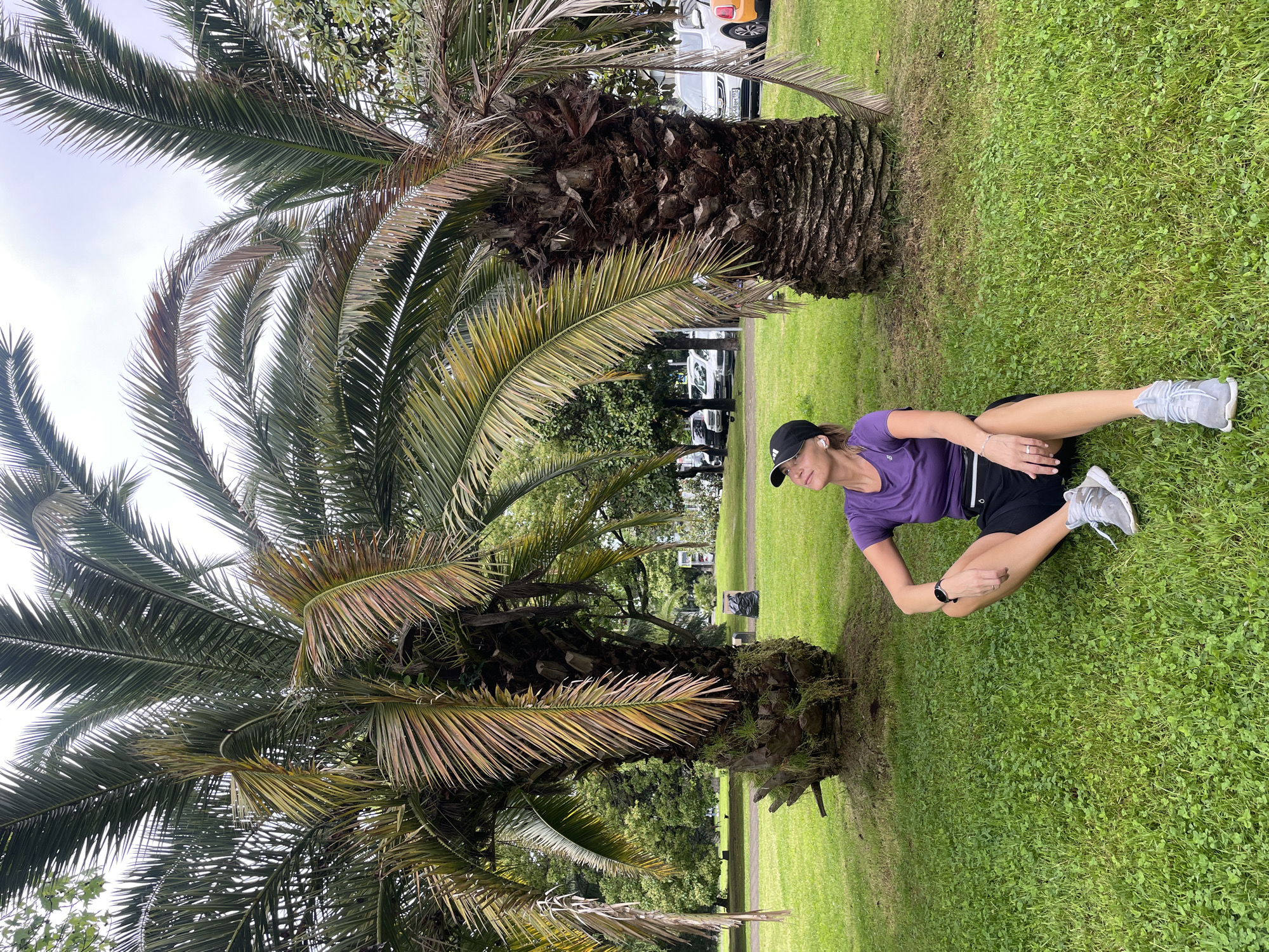 The image shows a woman sitting on the grass, stretching her leg, in what appears to be a park setting in Sochi, Russia. The location is identifiable by the presence of palm trees common to the Black Sea coast of Russia. The woman is wearing a purple shirt, black shorts, a black baseball cap, and grey sneakers. She is sitting with one leg extended and appears to be holding or stretching her foot. The background features green grass, several palm trees, other trees, and possibly parked cars. The sky appears overcast.