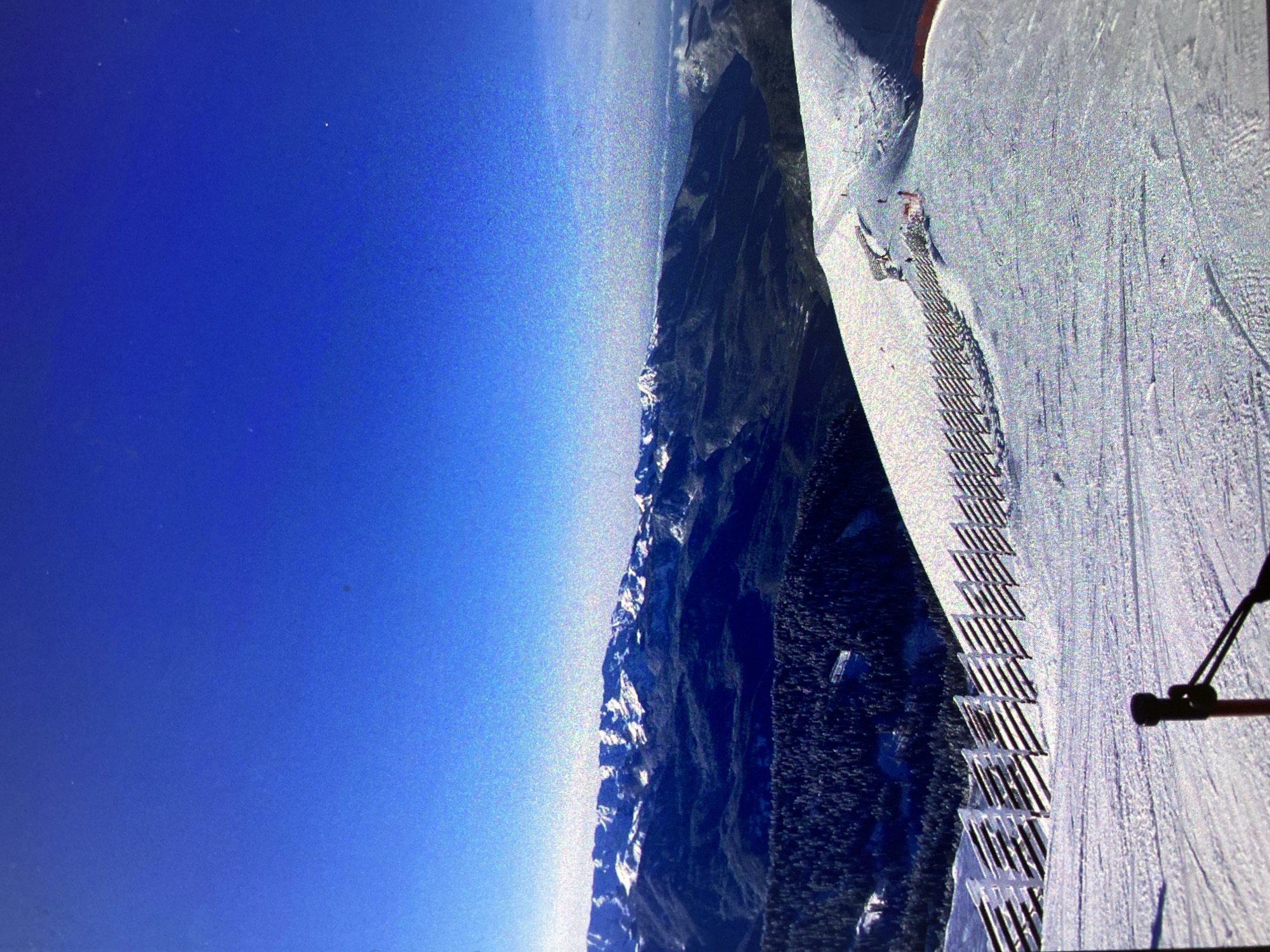 This image captures a expansive view of a snow-covered mountain landscape under a clear, bright blue sky, presented as if rotated 90 degrees clockwise. When correctly oriented, the vast blue sky fills the upper and left portions of the frame, showing a subtle gradient of color.
Along the horizon in the middle ground, a majestic mountain range unfolds with dark, jagged peaks and slopes partially covered in snow. Below these peaks, dense dark green forests can be seen, hinting at valleys or lower elevations.
In the foreground and extending towards the right, a wide, groomed ski slope dominates the scene. The snow on the slope exhibits distinct parallel grooves, indicating it has been recently prepared. A prominent feature on this slope is a series of grid-like barriers or fences, possibly for safety or avalanche management, running diagonally downwards. A small, distant figure, likely a skier or snowboarder, is visible on the upper section of the slope, suggesting active use of the area for winter sports. On the far right edge of the frame, a dark pole or structure, possibly part of a camera setup or ski lift mechanism, is partially visible.
The scene is bathed in bright daylight, with no clouds in the sky, indicating excellent, clear weather conditions. The overall impression is one of a vast, tranquil, yet active mountain ski resort. No visible text is present in the image.
While the provided location context is Saint Petersburg, Russia, the geographical features depicted—high, snow-capped mountains and extensive ski resort infrastructure—are more characteristic of mountainous regions like the Caucasus or Ural Mountains, rather than the relatively flat terrain around Saint Petersburg. It is likely that this image is a view displayed or transmitted from a location in Saint Petersburg, rather than being a depiction of the city itself.