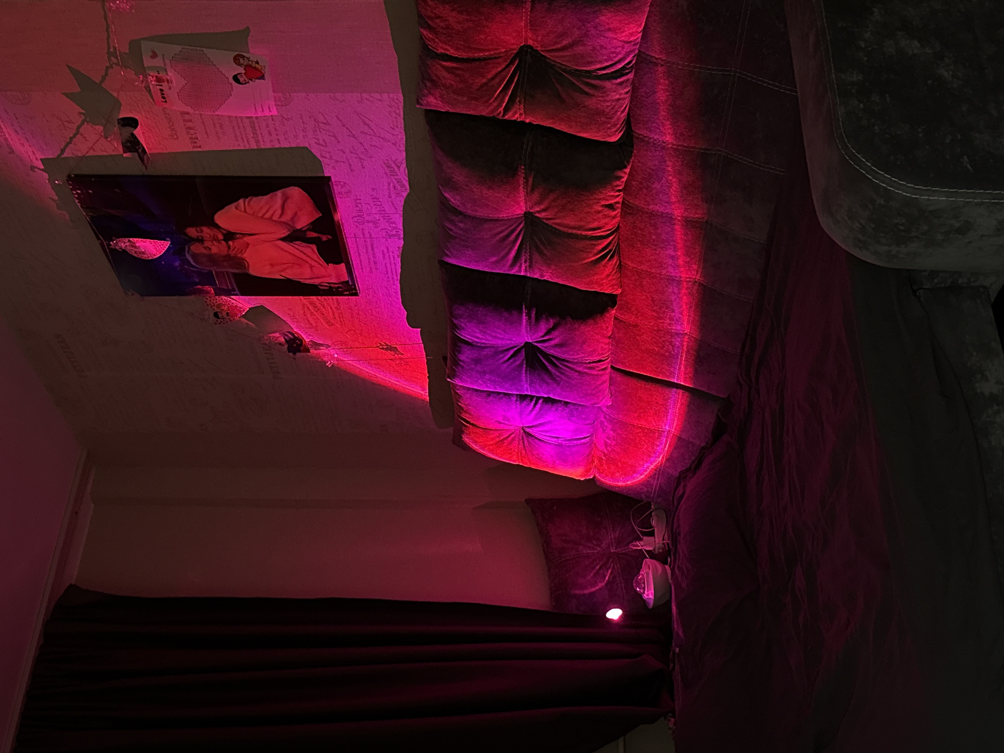 The image shows a room interior in Chaikovsky, Russia. The ceiling has a photo frame and a greeting card attached. A dark-colored curtain is visible at the bottom. A tufted sofa runs diagonally across the image, illuminated with red and purple light. Part of an armchair is seen on the right. The general lighting in the room is dim, with a red/purple hue.