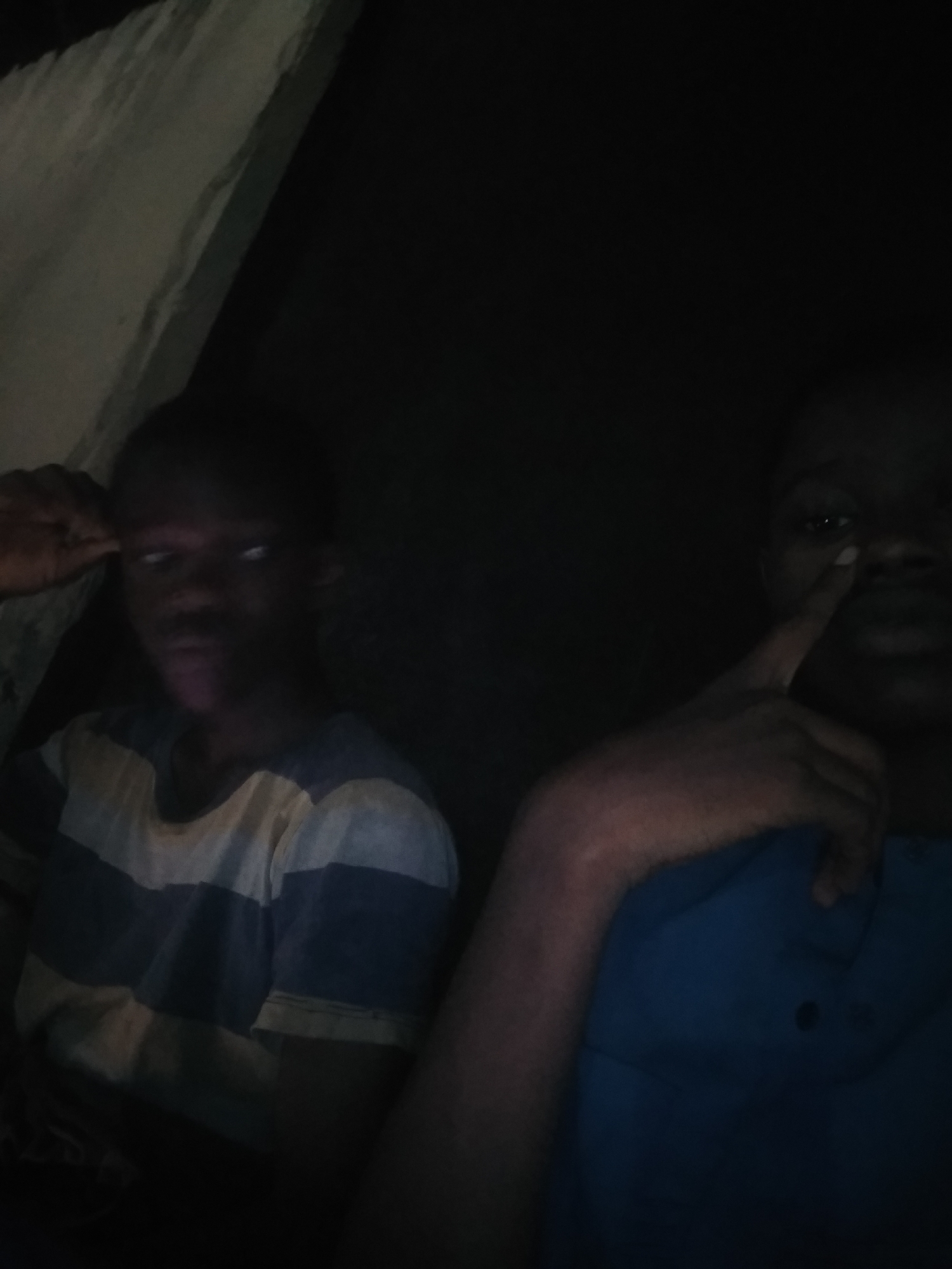 Two young men are in a dimly lit space. The man on the left is wearing a horizontally striped shirt, while the man on the right is wearing a dark blue shirt. The man on the left is resting his head on his hand, while the man on the right has his finger touching his nose. The location appears to be indoors and may be in Shangisha, Nigeria.