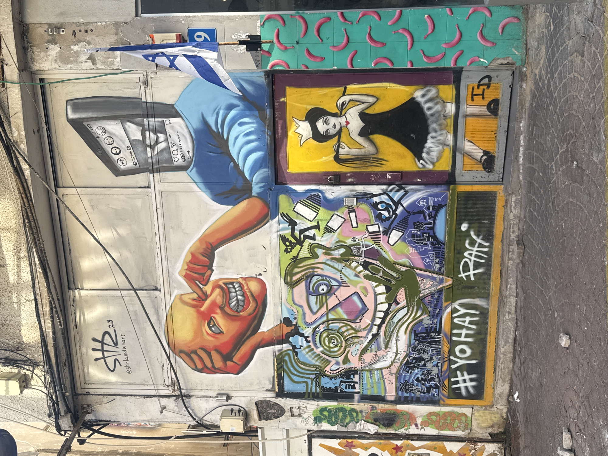 This media file captures a vibrant street art mural in Tel Aviv, Israel. The artwork is displayed on a metal surface, likely a garage door or shutters, which has been painted over multiple times. Dominating the composition is a large depiction of a person's head, rendered in a stylized and almost cartoonish manner with an orange hue and an exaggerated, wide grin. A hand, also orange, is shown poking the cheek of this head, creating a sense of playful interaction or perhaps a critique.
Above this central figure, a blue-sleeved arm extends from the top of the frame, holding what appears to be a stylized electronic device, possibly a vintage TV or monitor. On the monitor's screen, a face is drawn with several small circular icons, suggesting emotions or settings. A small Israeli flag is also visible, attached to a pole above the artwork.
To the right of the main figures, the mural continues with distinct panels. One panel features a black-and-white illustration of a woman wearing a crown and a black dress, with bright yellow background. Another panel above this is decorated with a repeating pattern of pink banana-like shapes on a teal background. Below the central head, a more abstract and chaotic piece of art unfolds, with a jumble of colors and shapes that create a complex facial or abstract design.
Visible text includes "#YOHAY PAFF" in white spray paint on a dark background, and "@shirlamdanart 23", possibly an artist's signature and year. The overall scene suggests an urban environment, with visible electrical wires and a cobblestone sidewalk on the right edge of the frame. The artwork is colorful and energetic, conveying a sense of urban creativity and commentary.