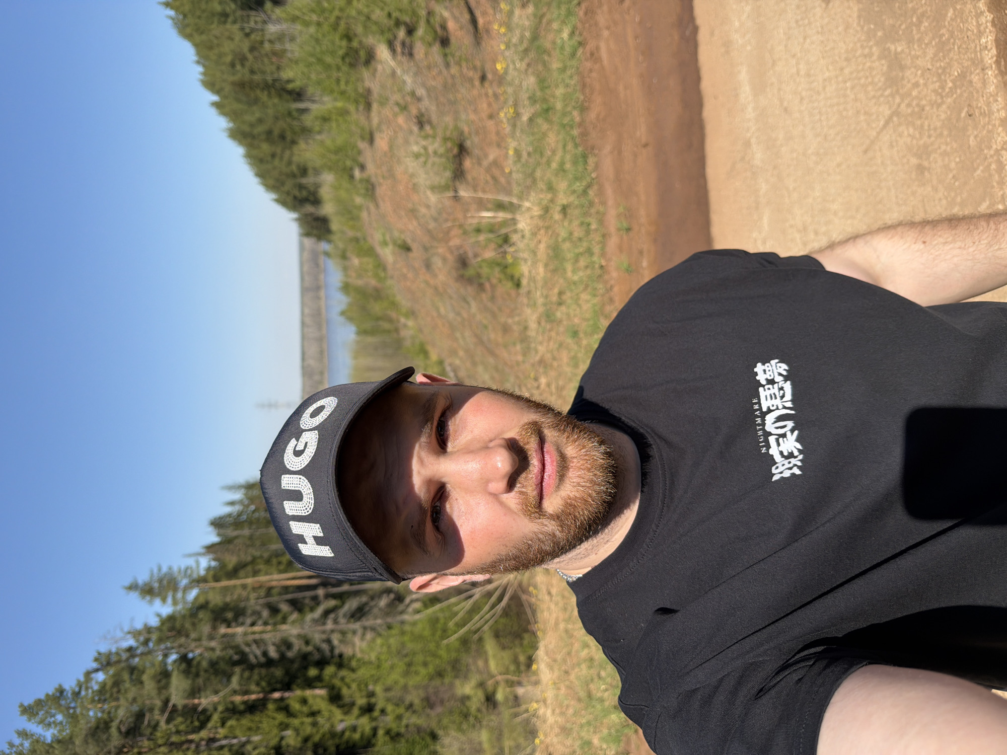 The image is a selfie of a man with a beard and a "HUGO" branded baseball cap, taken outdoors on a sunny day. The man is wearing a black t-shirt with Japanese text on it. The background shows a dirt path, trees, and a body of water in the distance. Based on the prompt, the location is likely Прикамский, Russia.