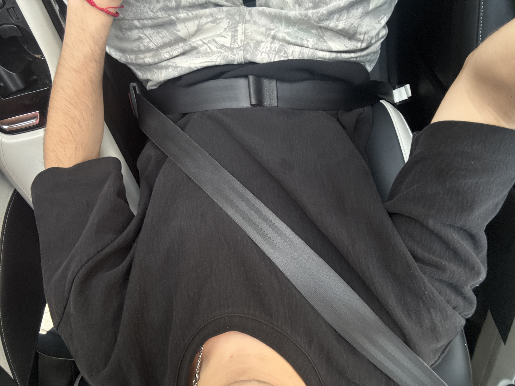 A person is sitting in the driver's seat of a car, wearing a seatbelt. The person is wearing a gray and white patterned shirt, a black shirt, and a silver chain necklace. The interior of the car is visible, with white seats and black dashboard elements. The image was likely taken in a car in San Bernardino Tlaxcalancingo, Mexico.