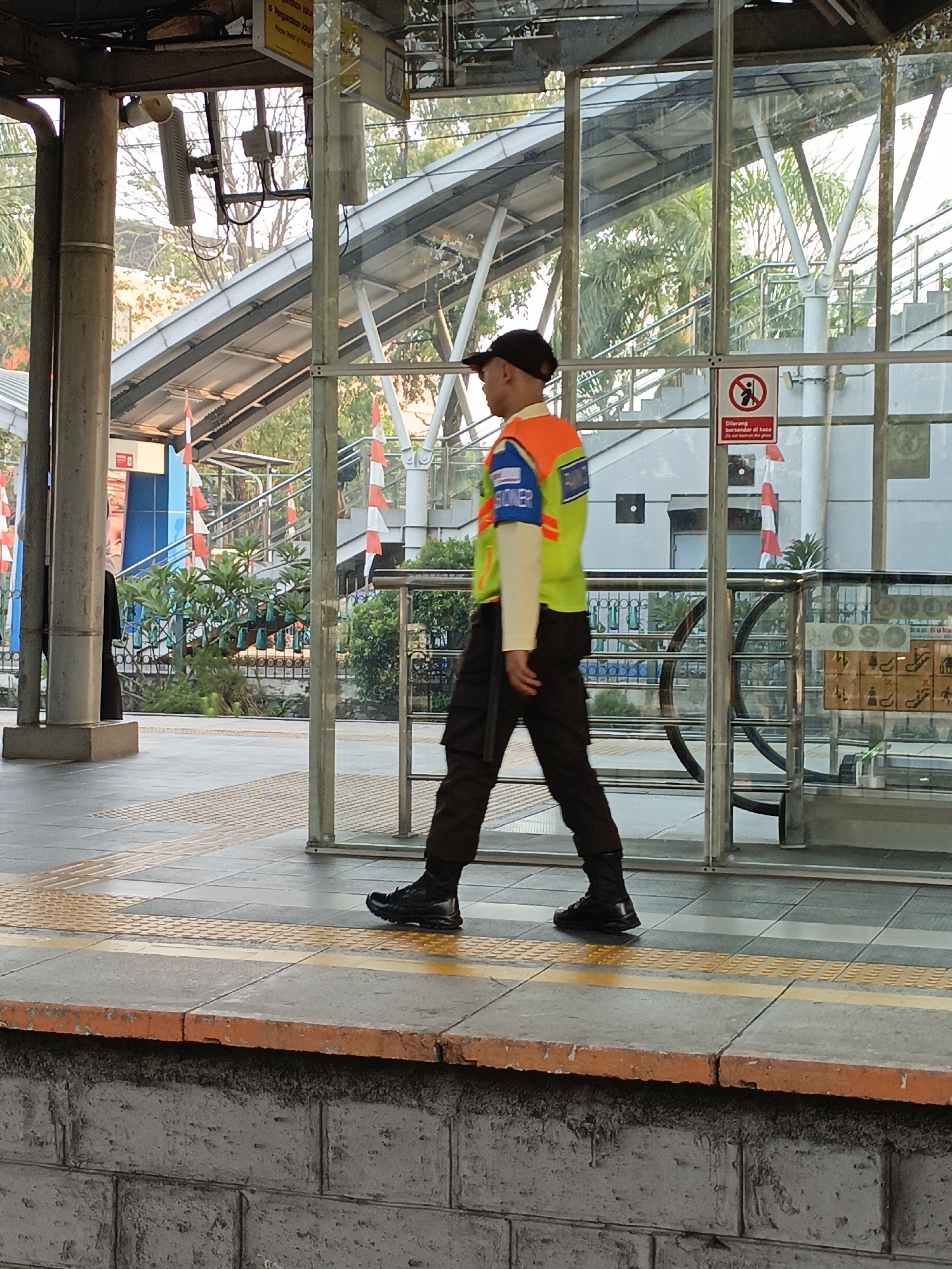 A person wearing a black baseball cap, a long-sleeved cream shirt, a bright orange vest over the shirt, and dark cargo pants walks on a tiled platform. The vest has blue and white patches on the chest and a blue and white patch on the left sleeve that reads "SPOONER". The person is wearing black work boots. The platform is bordered by an orange-painted concrete edge and a grey stone brick wall. In the background, there is a glass enclosure with metal framing. Inside the enclosure, a staircase with metal railings and white risers is visible, along with greenery and trees. Flags with alternating red and white stripes are displayed on poles outside the glass. A sign with a red circle and a diagonal line through a silhouette of a person is posted on the glass, along with text in Indonesian and English that reads "Dilarang bersandar di kaca Do not lean on the glass". Further into the background, there are metal structures and what appears to be an escalator or another staircase. The scene appears to be an outdoor public transit station, likely a train or bus station, in East Jakarta, Indonesia, given the Indonesian signage. The lighting suggests it is daytime.