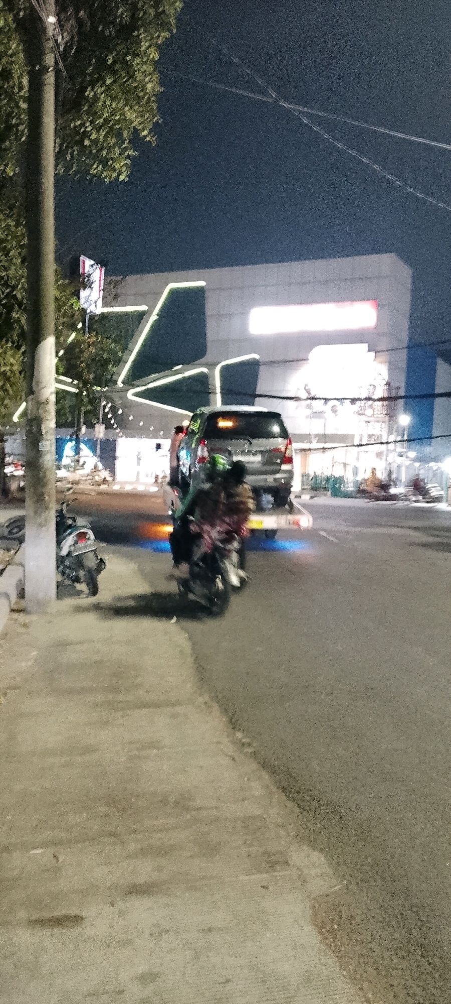 A nighttime, low-angle, vertical shot captures a street scene in Bekasi, Indonesia. A concrete sidewalk runs along the left side of the frame, bordered by a large tree and a utility pole. Across the asphalt road, a modern building with geometric accents and neon lighting stands prominently. A silver SUV is in the mid-ground, its brake lights illuminated. In the foreground and mid-ground, two motorcycles are visible, one parked near the sidewalk and another in motion on the road, carrying two occupants. The road surface appears wet, reflecting the light from the vehicles and the building. The sky is dark, indicating the late hour.
