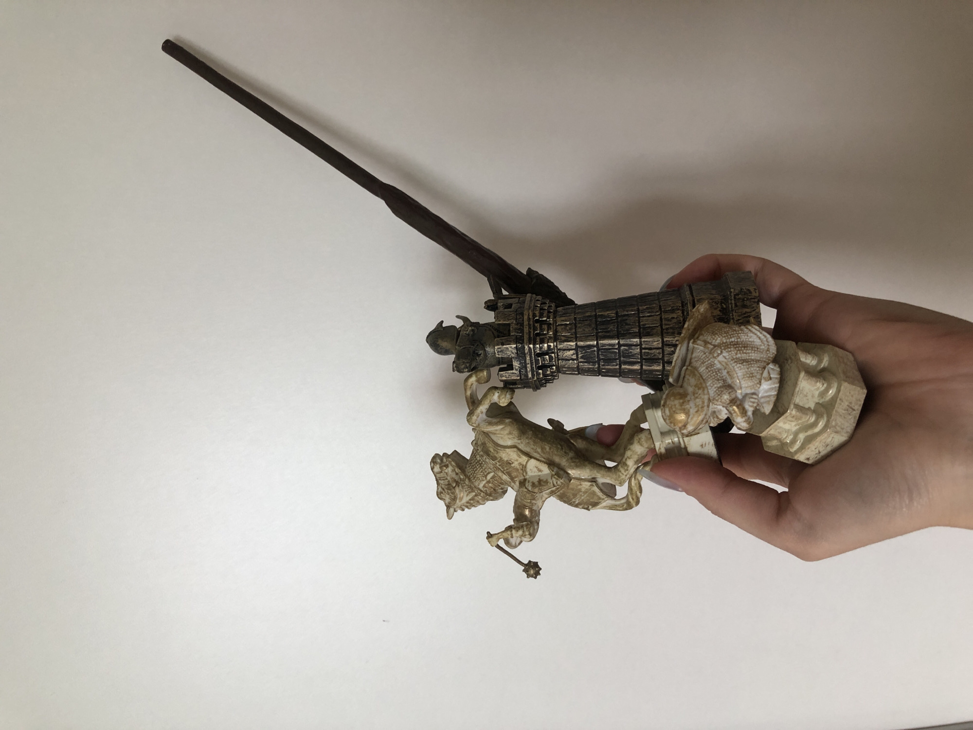 A hand holds a decorative tower figurine, brown with a dark brown rod extending from its top. The figurine includes sculpted figures of animals. The background is a plain white surface.