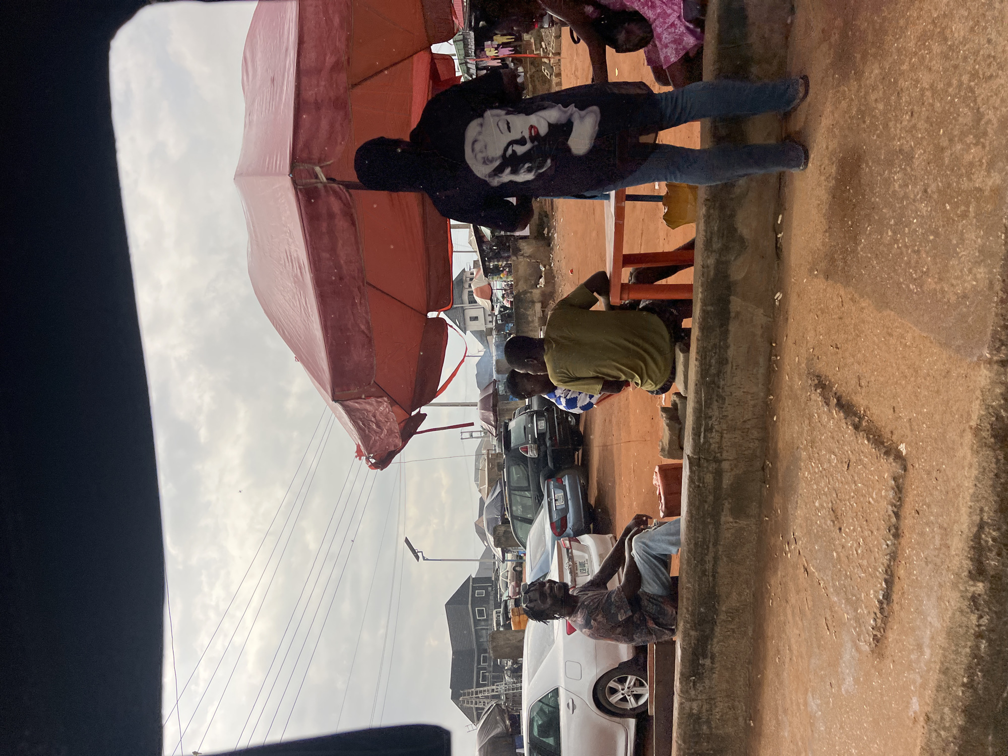This image depicts an outdoor market scene in Okpanam, Nigeria, under a cloudy sky. In the foreground, a man with dreadlocks and a patterned t-shirt sits on a concrete ledge, smiling at the camera. He is wearing blue jeans. To his right, a person in an olive-green t-shirt is seated. Further to the right, a person wearing a black t-shirt with a large image of Marilyn Monroe on the back stands near an orange plastic table. A person in a pink outfit is also visible to the far right.
Behind the seated individuals, several parked cars are visible, including a silver sedan, a dark grey SUV, and a blue sedan. The background shows various market stalls and buildings, some with umbrellas providing shade. Power lines stretch across the sky. The ground is a reddish-brown earth. The overall impression is of a bustling, informal marketplace.