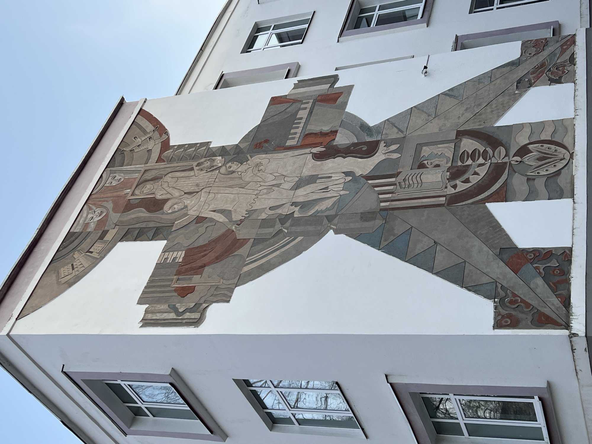 The image shows a mural on the side of a building in Chaikovsky, Russia. The mural is painted in a stylized, geometric Art Deco style, with a muted color palette of browns, grays, and whites. It depicts a complex arrangement of figures, objects, and symbols including human figures lying down, musical instruments (piano and violin), and abstract designs. The building is white with a light-colored trim around the windows. There are square windows above and below the mural. The sky is light blue. The camera angle is looking upwards.