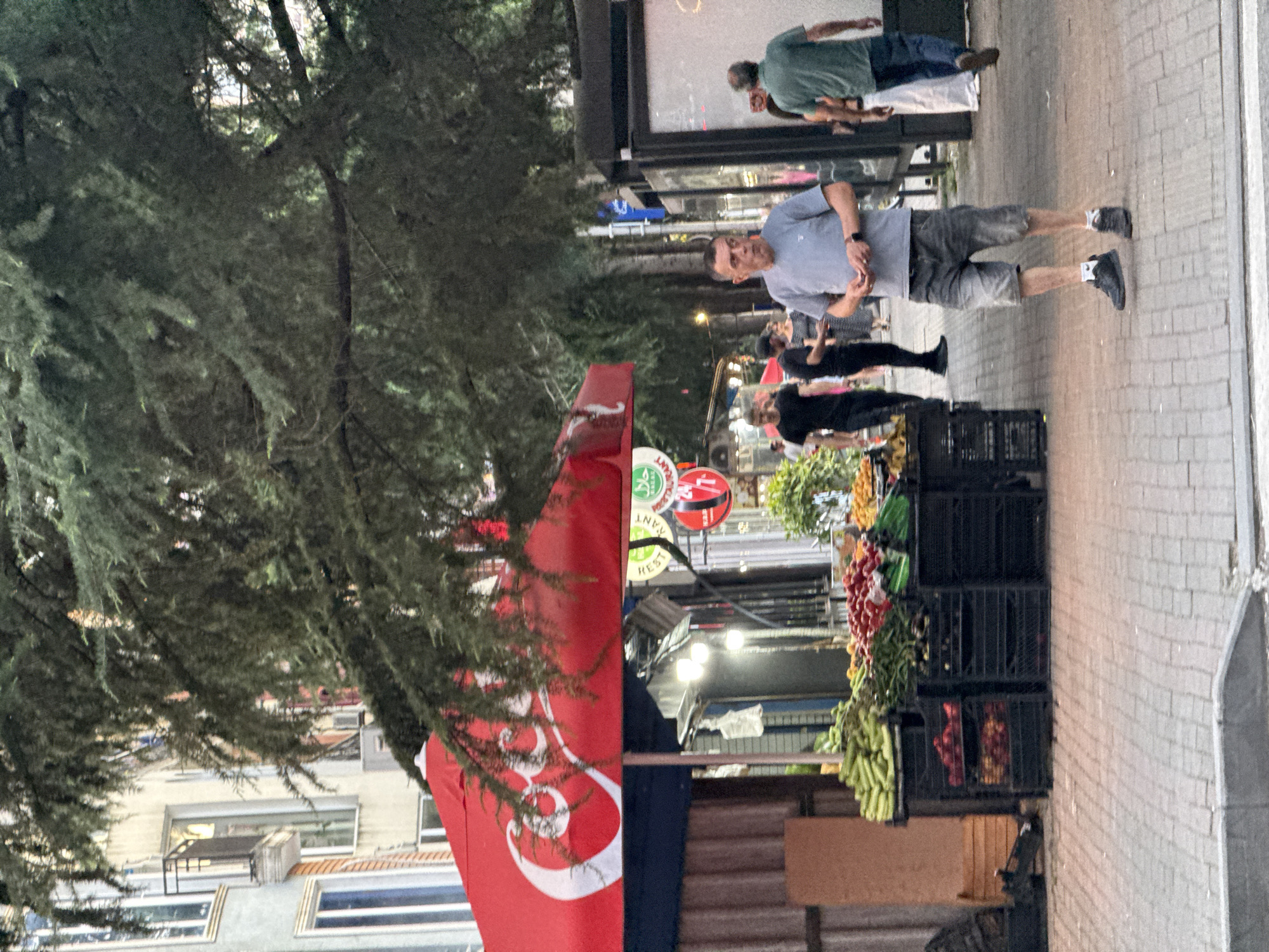 An outdoor fruit stand is depicted under a large Coca-Cola umbrella. The stand displays an assortment of fresh produce, including cucumbers, tomatoes, and other vegetables, arranged in black crates. Several individuals are visible on a paved sidewalk. A man in a gray t-shirt and denim shorts stands in the foreground, looking to his left. Behind him, other people are walking. The background shows a building with balconies and windows, and a tree with dense foliage arches over the scene. The lighting suggests it is daytime.
