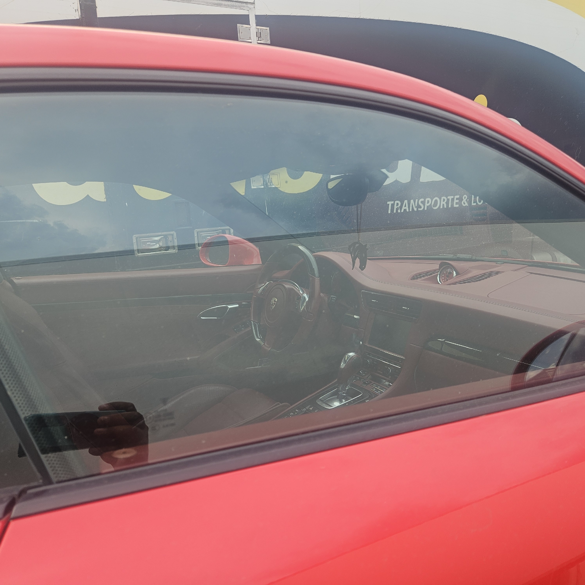 The image shows the interior of a red car, specifically the dashboard, steering wheel, and part of the driver's seat, viewed through the passenger window. A person's hand is visible holding a dark-colored smartphone, reflecting in the glass. The car appears to be parked, with the background showing a building with yellow lettering that reads "TRANSPORTE & LOG." This is consistent with the location of La Jonquera, Spain, which is known for its border crossing and logistics facilities. The interior of the car is detailed, with brown leather seats and a sporty steering wheel. The lighting suggests it is daytime.