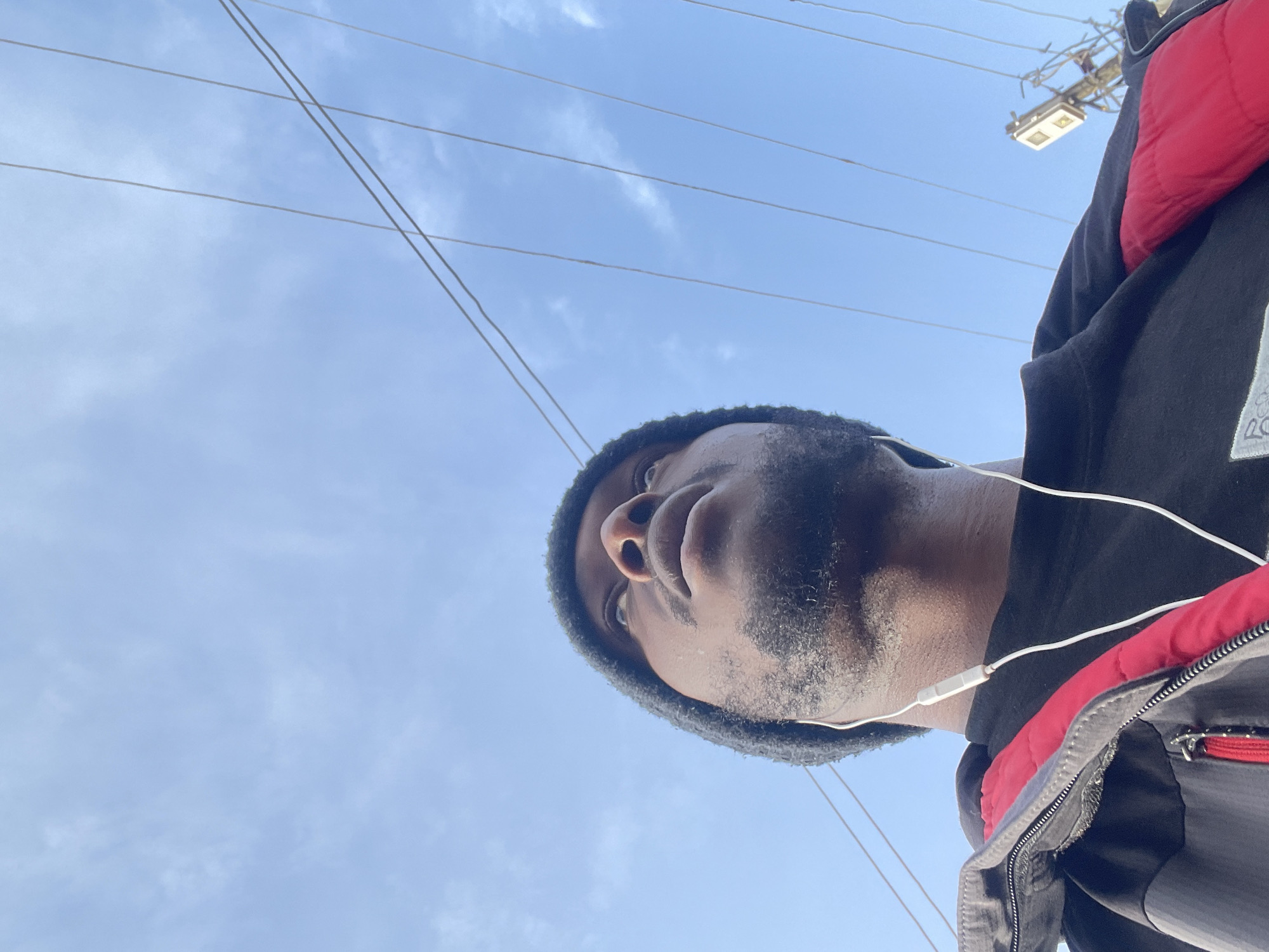 An outdoor, low-angle, medium shot captures a Black man wearing a dark beanie, black t-shirt, and a black and red jacket. He is looking upward, and white earbuds are visible. The background consists of a clear blue sky with thin clouds and several power lines stretching across it. In the far right background, a utility pole with electrical equipment is visible. The image is taken from a perspective that emphasizes the sky and the man's upward gaze. It is not possible to determine the specific location in Jos, Nigeria based on the visual information provided.
