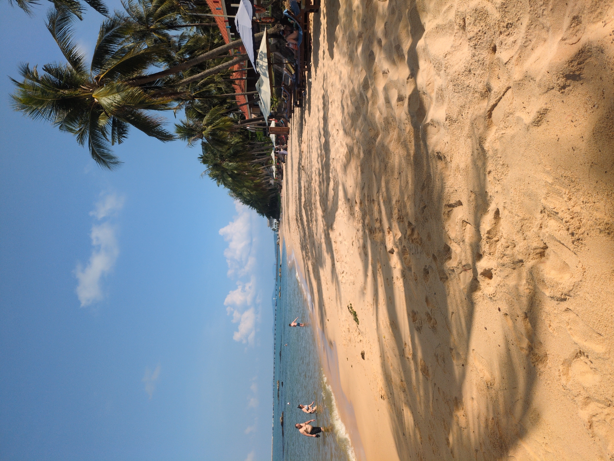 A beach in Phú Quốc, Vietnam, is pictured on a sunny day with a clear blue sky and scattered clouds. Palm trees line the edge of the beach, casting long shadows on the sand. People are wading in the shallow water along the shoreline, and umbrellas with chairs are arranged near the trees. The sand is light brown with visible textures.