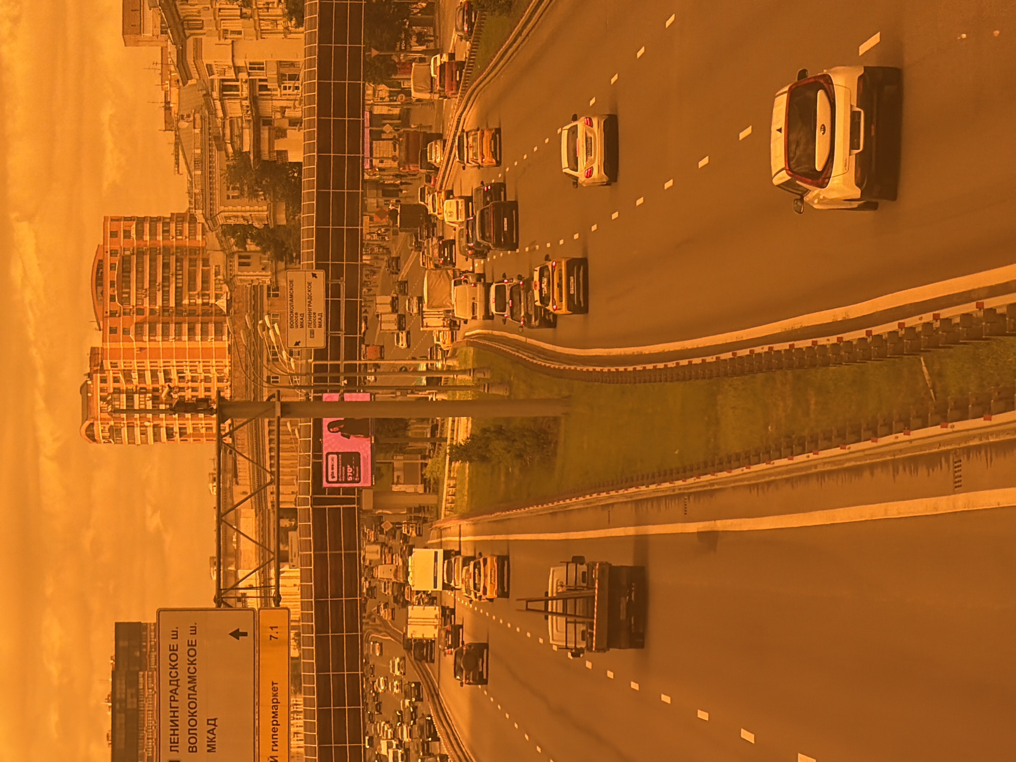 An aerial, medium shot captures a highway with moderate traffic under a bright, orange-tinted sky. The sky is a uniform, vibrant orange, obscuring any visible clouds or celestial bodies, and casting an overall warm, hazy glow on the scene. A multi-lane highway stretches diagonally from the bottom left to the top right of the frame. Cars, trucks, and vans are scattered across the lanes, with some vehicles appearing to be in motion. The road itself is asphalt with clear white lane markings. On the left side of the highway, a tall signpost displays directional information in Russian, with text such as "ЛЕНИНГРАДСКОЕ ш.", "ВОЛОКОЛАМСКОЕ ш.", and "МКАД". Another sign below indicates "и гипермаркет" with a distance of "7.1". To the right of the highway, a grassy verge and a barrier are visible. In the background, multi-story buildings, including apartment complexes and older architectural structures, rise against the orange sky. A pedestrian overpass crosses the highway in the mid-ground. An advertising billboard on the right side of the highway displays an image of a woman and text including "570". The overall impression is a busy urban highway scene under an unusual, intensely orange sky.