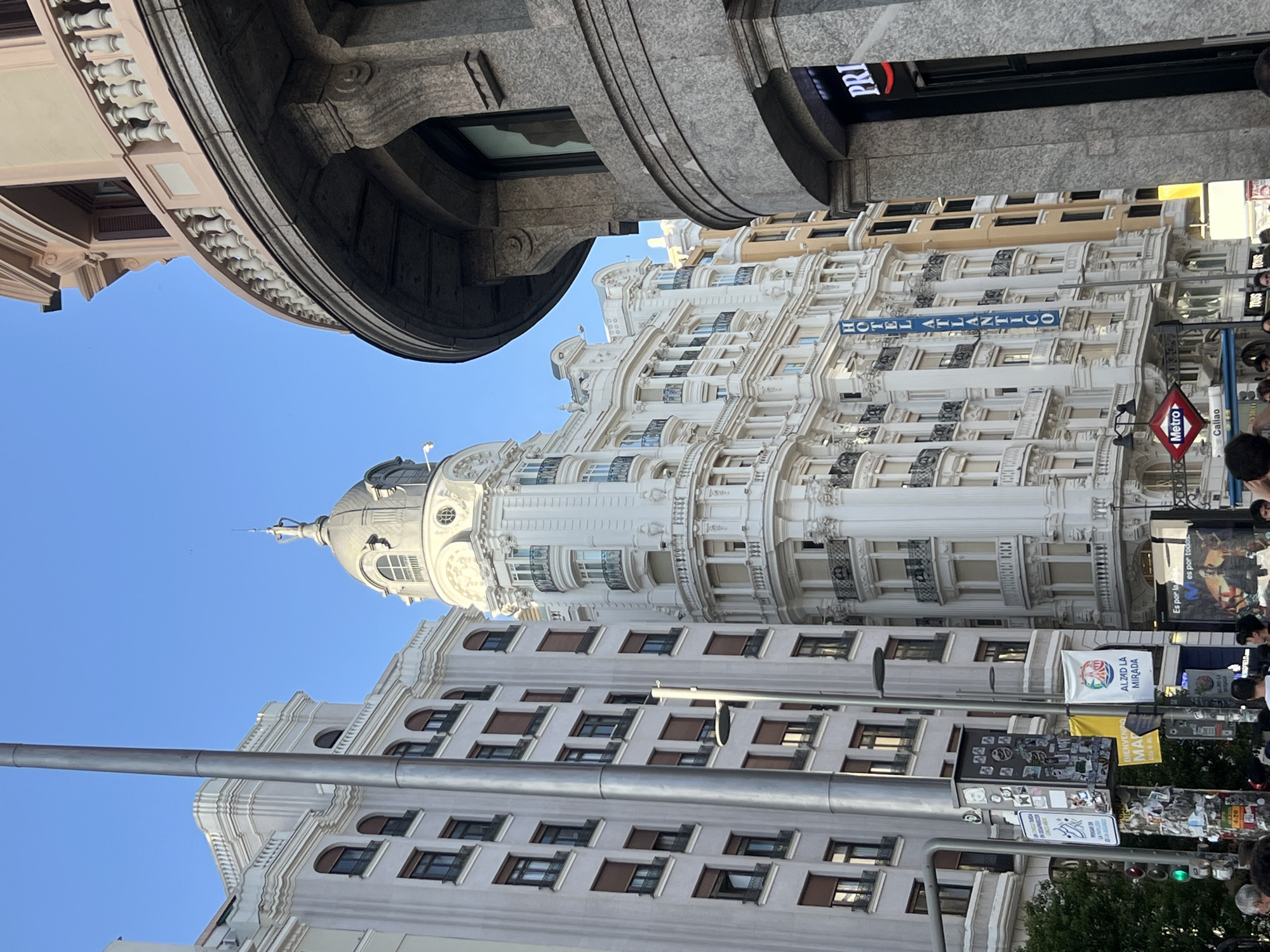 The image shows a view of the intersection of Gran Vía and Calle de Preciados in Madrid, Spain. Visible landmarks include the Hotel Atlántico with its distinctive dome, a Metro sign indicating the Callao station, and a commercial building featuring the "PRI" sign. There is a crowd on the street and various advertisements, signs, and traffic lights are present. The buildings are ornate with classical architectural details, and the sky is clear.