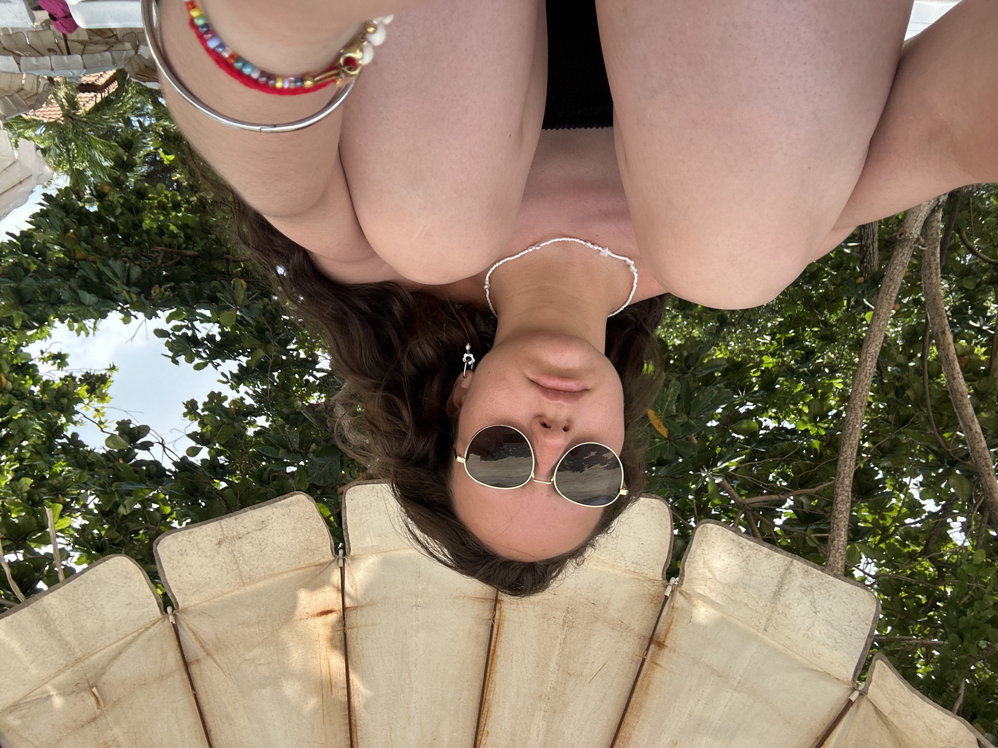 From a low-angle perspective, a woman in black swimwear poses under a parasol on a beach in Ao Hin Khok, Thailand. She wears round sunglasses, a beaded necklace, and bracelets on her left wrist. Her brown hair cascades down. In the background are trees and a glimpse of the sky. The parasol is cream-colored and constructed of panels.