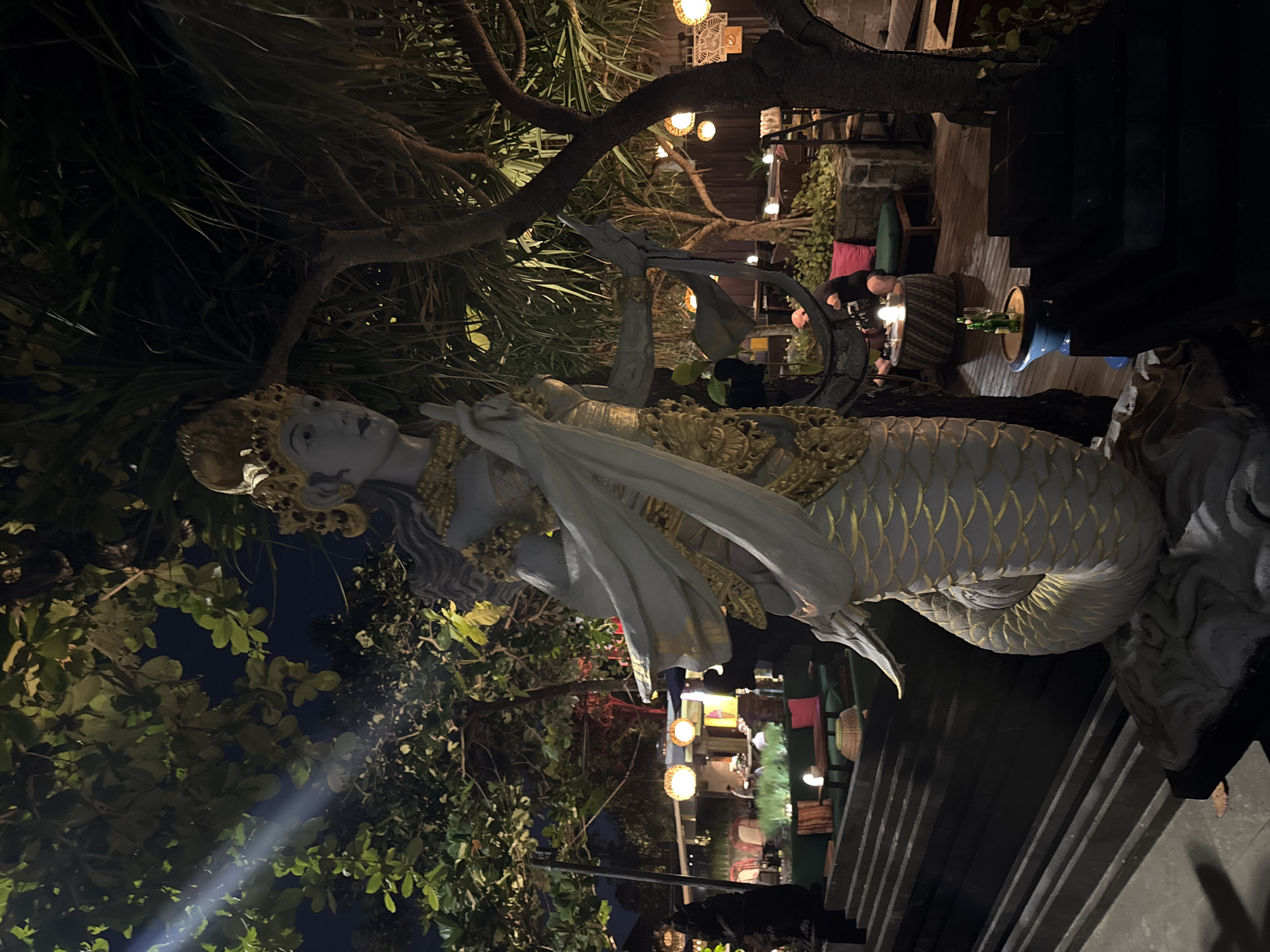The image depicts a night scene at a restaurant or bar in Kuta, Indonesia, likely near the beach. In the foreground, a large statue of a mermaid-like figure with a human torso and a serpentine lower body stands prominently. The statue is white with gold accents on its headdress, chest, and scales. It is illuminated by artificial lighting, creating a dramatic effect.
Behind the statue, a multi-tiered seating area is visible, with lush green foliage and trees providing a natural backdrop. Several round, illuminated lanterns are suspended from the trees, casting a warm glow. The seating area is furnished with dark-colored tables and chairs, and some areas are set up with cushions in shades of green and pink.
In the middle ground, several patrons are seated at tables, some appearing to be dining or socializing. The lighting is dim, creating an intimate atmosphere. A person is seated at a round table with a candle, and another table has two bottles of sparkling wine and two glasses. The overall environment suggests a relaxed, outdoor dining or entertainment venue. The architectural style of the area appears to incorporate natural materials and possibly Balinese influences.