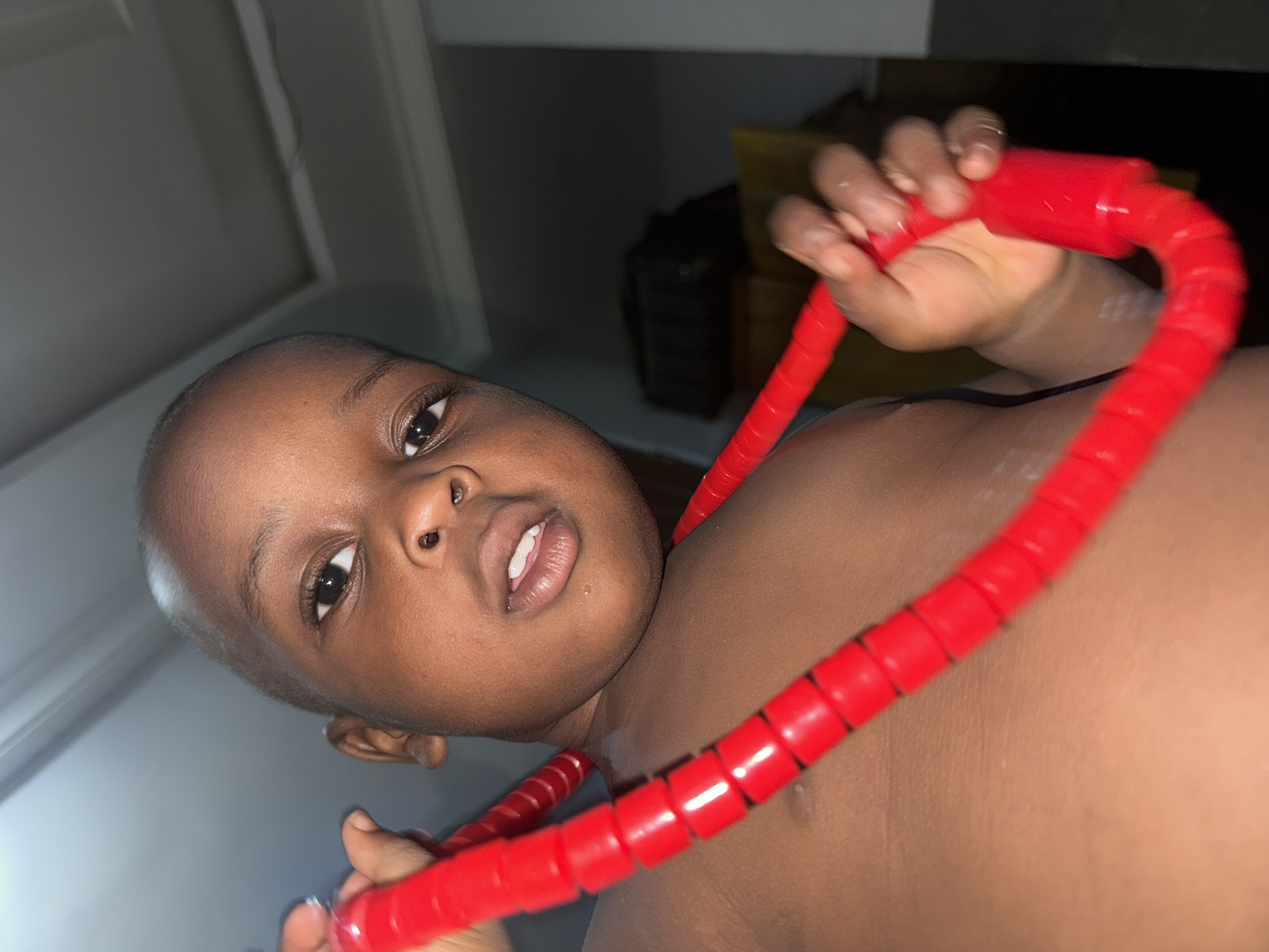 A close-up, slightly angled photograph shows a young Black child, likely between 2 and 4 years old, holding a bright red, segmented plastic tube. The tube is shaped in a semi-circle, extending from the child's right shoulder, across the chest and neck, and down to their left side, where it is held in their left hand. The child's head is positioned to the left, with their face looking towards the camera. The background appears to be an interior space, with a white wall visible in the upper left corner and some dark, undefined shapes and a yellow object suggesting furniture or storage behind the child. No indication of Sabon Tasha, Nigeria, is visible.
