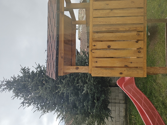 The image displays an outdoor children's play area located in Donchevo, Bulgaria, under an overcast sky. A wooden play structure occupies the right and center of the frame, comprising a raised platform with a vertical wooden plank railing and a gabled roof covered in dark red hexagonal asphalt shingles. A red plastic slide, featuring a subtle wave pattern, extends diagonally downwards from the left side of the play structure to the grassy ground. A grey concrete block wall is visible behind the slide. A large, dense evergreen tree stands prominently in the left background, with distant residential roofs partially visible on the horizon. The ground is covered in green grass. No individuals are present in the scene.