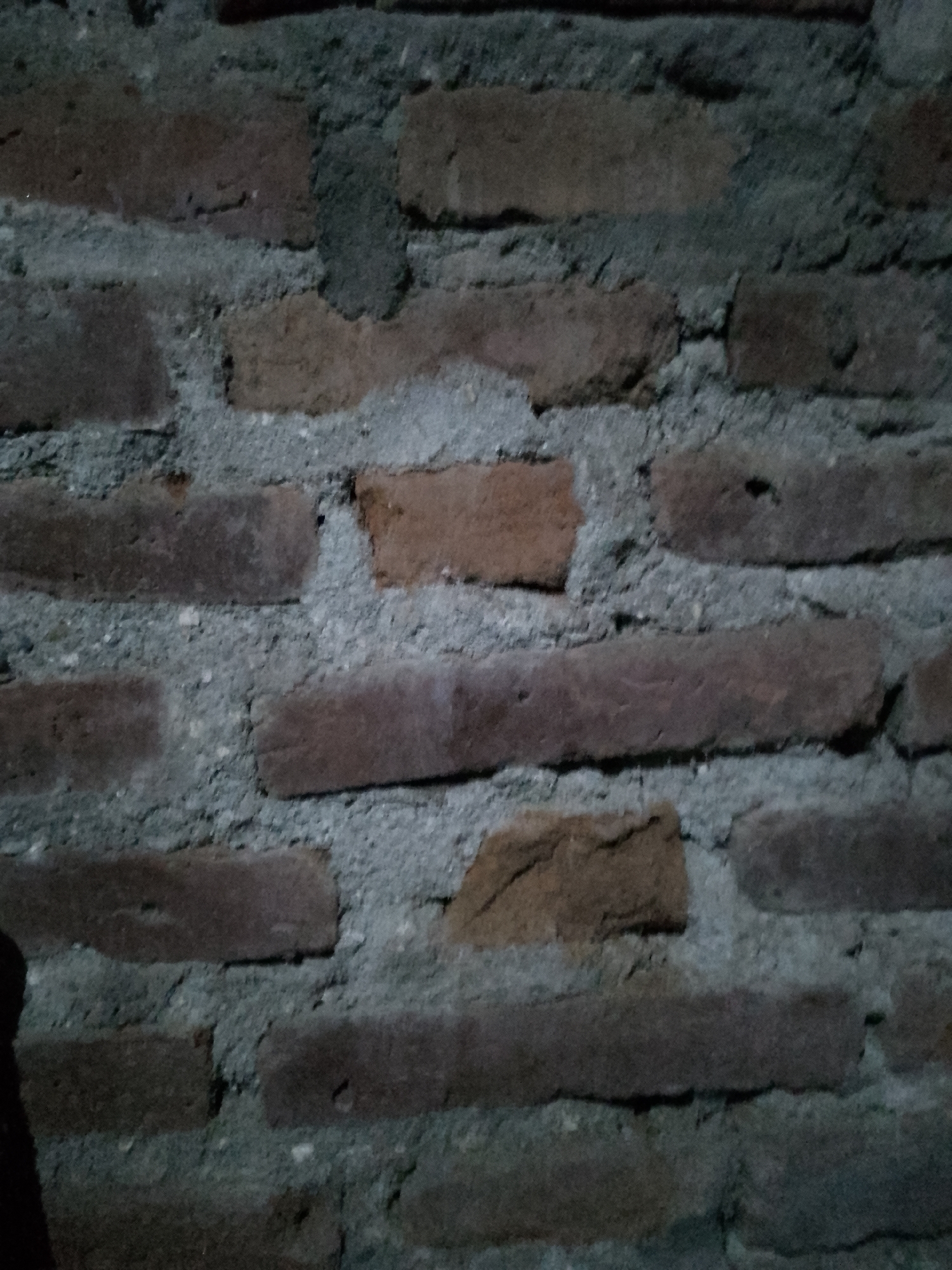 The image is a close-up, dimly lit photograph of a brick wall. The bricks are predominantly red and brown, with visible mortar in between, which appears to be grey and textured. The wall shows signs of age and weathering, with some bricks appearing uneven or chipped. There are no people or identifiable objects other than the wall itself. The setting appears to be outdoors or in an area with a rough, unfinished surface, consistent with a building exterior or interior wall. The specific location in Kauman, Indonesia, cannot be determined from the visual information provided in the image alone.