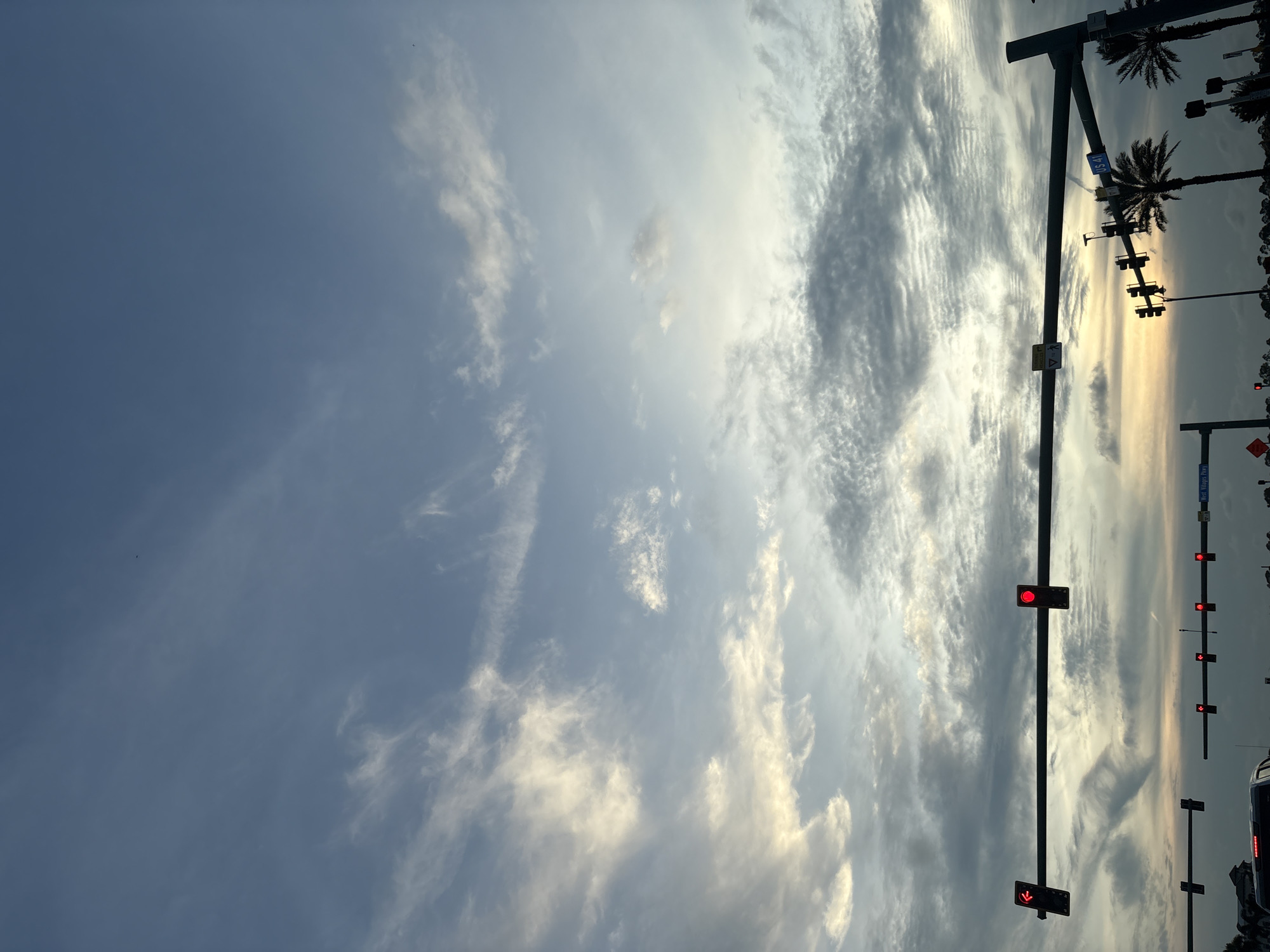 The image shows a view of the sky above a road in North Port, Florida, USA. The sky is filled with a mix of light and dark clouds, with some areas illuminated by the setting sun, creating a soft, warm glow. Several traffic lights are visible, with at least two showing red lights. Palm trees are silhouetted against the sky, indicating a tropical or subtropical location. A street sign reading "US 41" is also visible. The scene suggests a roadside view during twilight.