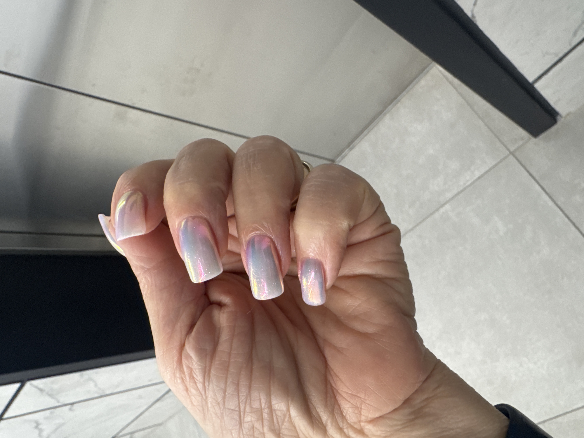 The image shows a close-up of a person's hand with acrylic nails. The nails are square-shaped and have a pearlescent, iridescent finish that shifts between pink, blue, and gold tones. The hand is resting against a tiled floor. The background includes a dark vertical element and a light-colored wall. The scene appears to be indoors, possibly in a public restroom or a salon. There is no discernible information in the image to place it specifically in Novosibirsk, Russia.