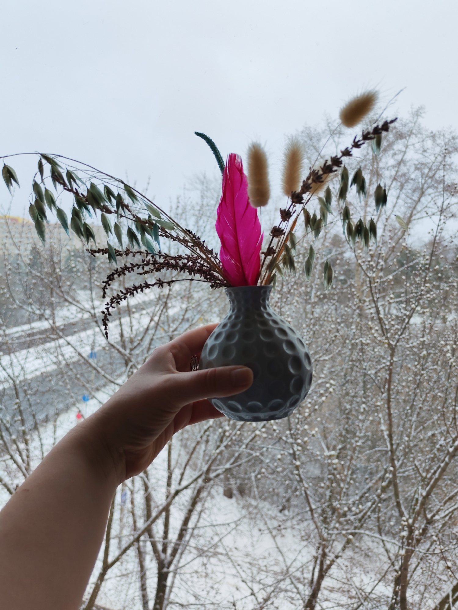 A person's hand is holding a small, gray vase filled with dried decorative plants and a bright pink feather. The vase is round with evenly spaced, spherical indentations. The dried plants include stems with seed pods, fluffy beige stalks, and a stem with small, dark purple flowers. The background is a blurred winter scene viewed through a window, showing trees covered in snow and indistinct buildings in the distance. The location is Izhevsk, Russia.