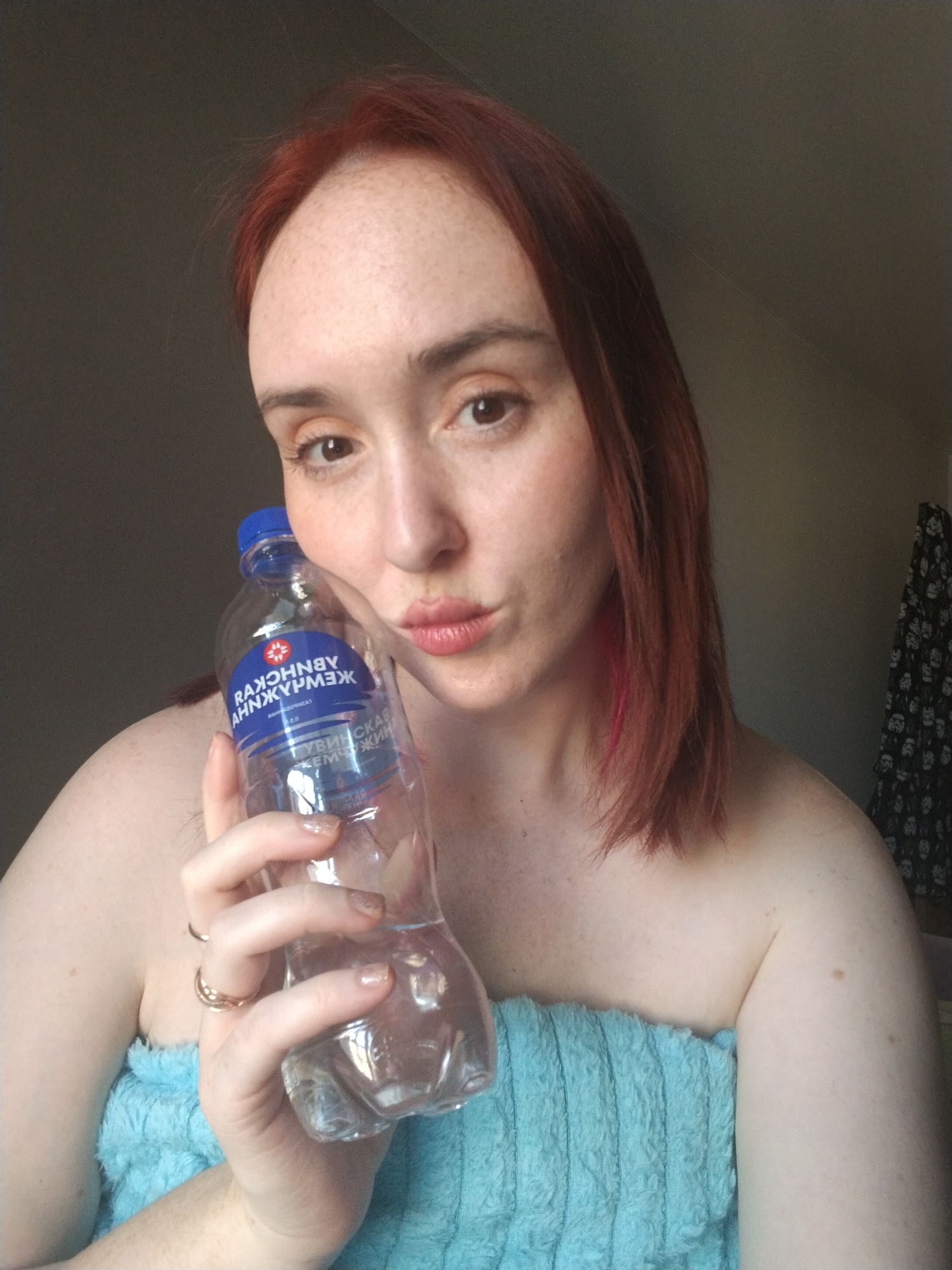 A person with light skin, reddish-brown hair with a pink streak, and freckles is holding a clear plastic water bottle. The person is wearing a light blue towel, possibly wrapped around their shoulders. They are pursing their lips as if giving a kiss. The bottle has a blue cap and a blue label with Cyrillic text on it, with "РАЖОННВУ АННЖУРМЭЖ" prominently displayed. The background is dimly lit, with a beige wall and a glimpse of a floral-patterned fabric hanging in the upper right corner. The image does not provide any identifiable information about the location being Izhevsk, Russia.
