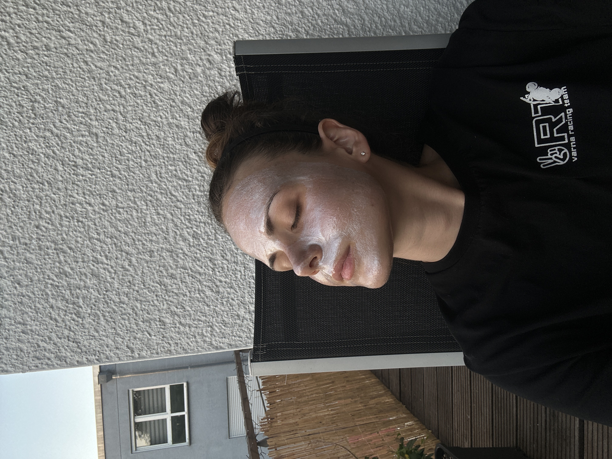 A woman is seen lying on a dark gray chair with her eyes closed. Her face is covered in a white cream, likely a face mask. She has her hair pulled back into a bun, and is wearing a black t-shirt that has "varna racing team" printed on it with a motorcycle graphic. The background shows a light gray building with a window, a textured white wall, and a wooden deck. Location: Unknown, Unknown.