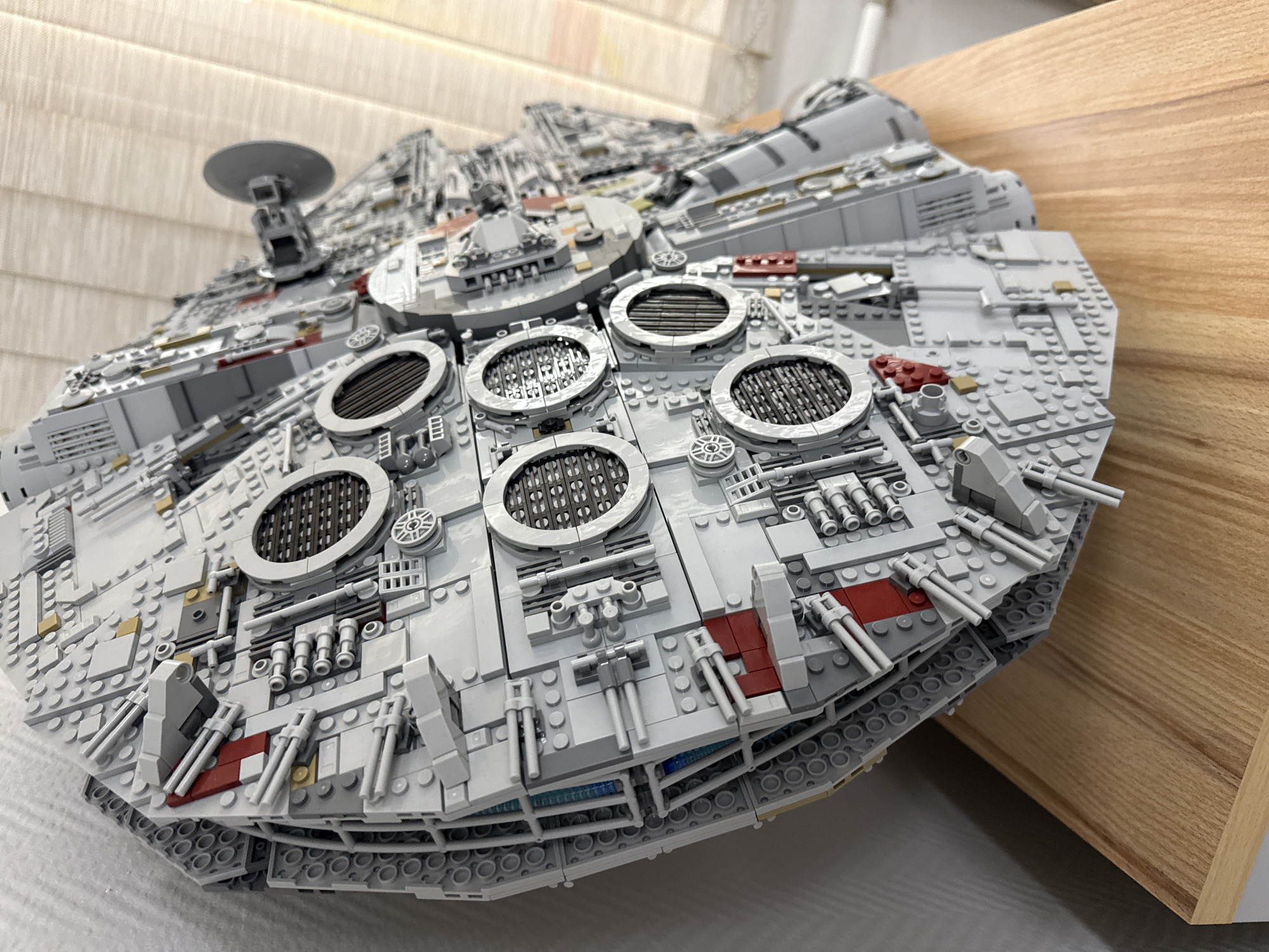 The image shows a detailed LEGO model of the Millennium Falcon. The model is predominantly gray and composed of numerous LEGO bricks. It's resting on a surface with a light-colored wooden texture, possibly a table or shelf. The background consists of a light-colored wall with vertical linear patterns. There is no indication in the image that places the scene in Krasnoyarsk, Russia. No people or actions are visible.