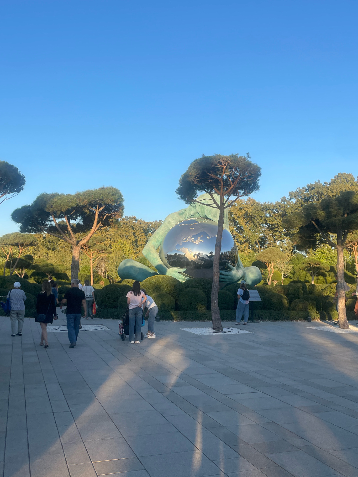 This image depicts a sunny day in the Park of Krasnodar, Russia, also known as "Galitsky Park." The park features a large, iconic sculpture of a hand holding a metallic sphere, reflecting the surrounding greenery and sky. Several people are present, walking along a paved pathway and admiring the artwork. The park's landscape is characterized by manicured bushes, carefully pruned trees, and patches of white gravel. The sky is clear and blue, indicating fair weather. The overall scene conveys a sense of a pleasant outdoor experience in a well-maintained public space.
