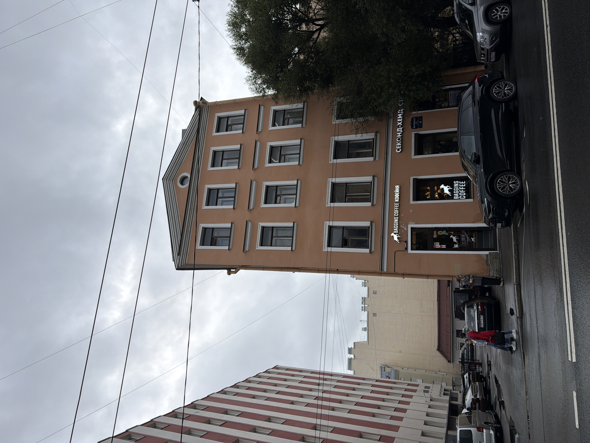 The image shows a street scene in Saint Petersburg, Russia, on a cloudy day. In the foreground, a black SUV is parked on a wet asphalt street. To the right of the SUV, a silver SUV is also parked. Further back, a multi-story orange building with several windows is visible. Above the ground floor, there is a sign that reads "BAGGINS COFFEE" with a silhouette of a leaping goat. To the right of the coffee shop sign, another sign reads "СЕКОНД-ХЕНД". A few people are visible on the sidewalk, and cars are parked along the street. To the left of the orange building, a taller building with red and white stripes is partially visible. The sky is overcast with clouds, and utility lines are strung across the frame.