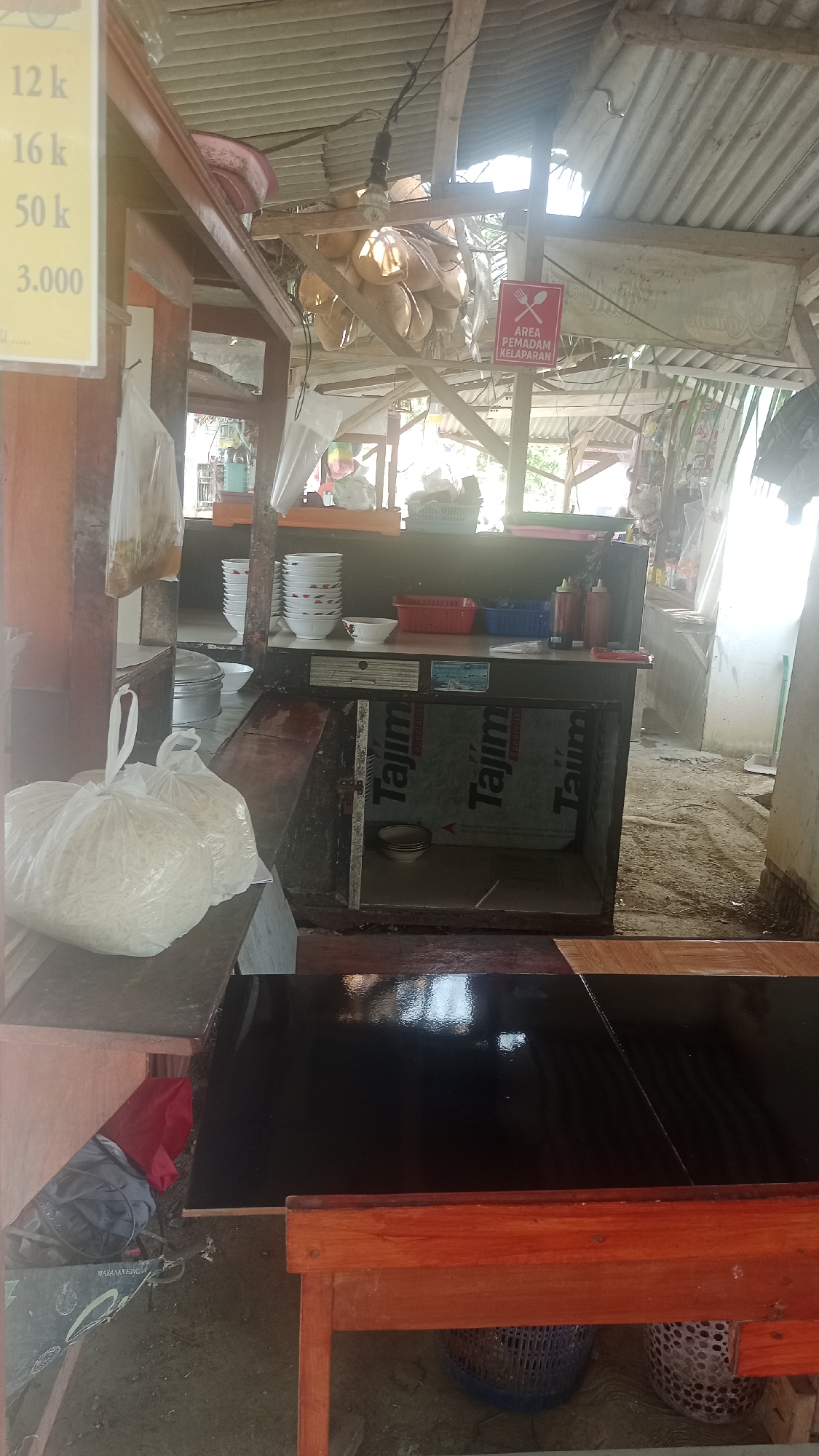 The image shows an outdoor food stall in Cibeber, Indonesia. A stall owner is partially visible behind the counter. The stall is made of wood and corrugated metal. There are several bags of noodles and bowls on display. A sign in red reads "AREA PEMADAM KELEPAKAN," which translates to "Hunger Extinguisher Area." The ground is dirt and debris.
