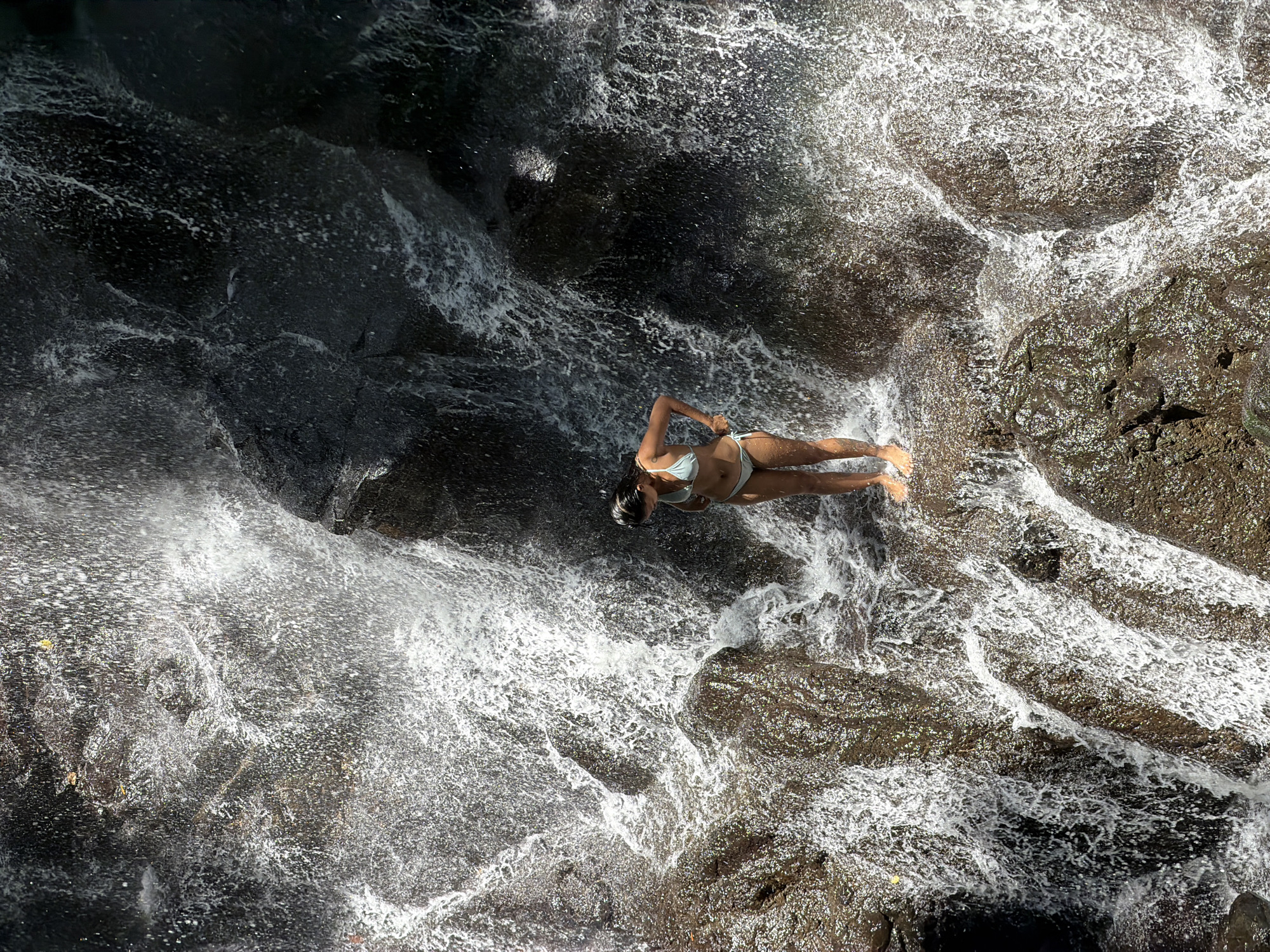 A high-angle shot depicts a woman in a white bikini lying on a rock formation amidst a waterfall. Her body is positioned diagonally across the frame. Her head is in the upper part of the frame with her feet pointing to the lower part. Water cascades around her and the rocks. The water is frothy and white where it is turbulent, and darker where it is deep. The rocks are dark, with some showing signs of moisture. The location is the city of Beng, Indonesia.