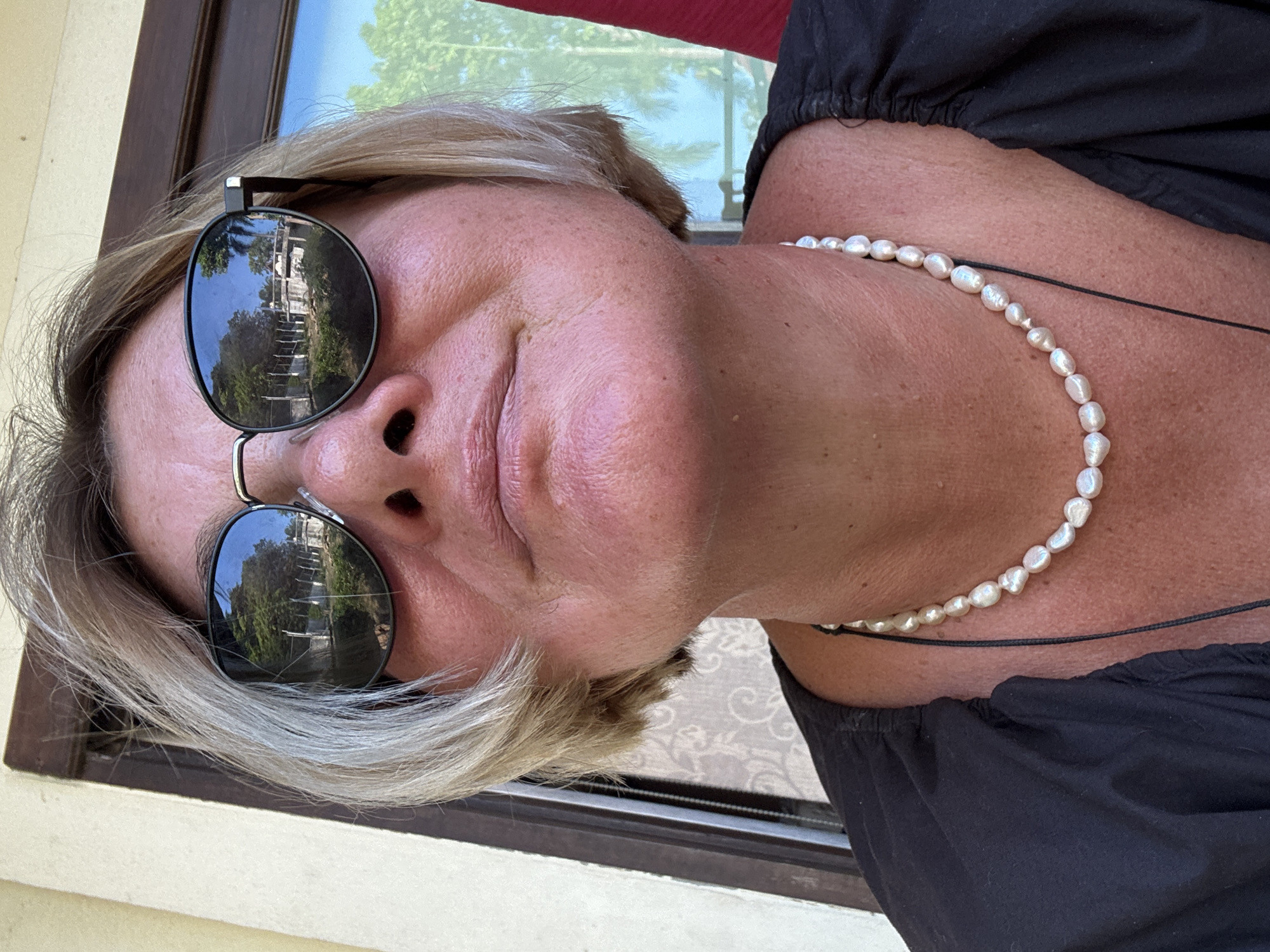 A woman with short blonde hair is shown from the chest up, wearing sunglasses that reflect an outdoor scene. She has a pearl necklace on a thin black cord around her neck and is wearing a dark-colored, off-the-shoulder shirt. The scene reflected in her sunglasses appears to be stairs or a pathway leading upward, surrounded by trees and greenery. A building is visible at the top of the stairs. A partial view of a window frame and a light-colored wall are in the background. The location is บ้านบางรักษ์, Thailand.