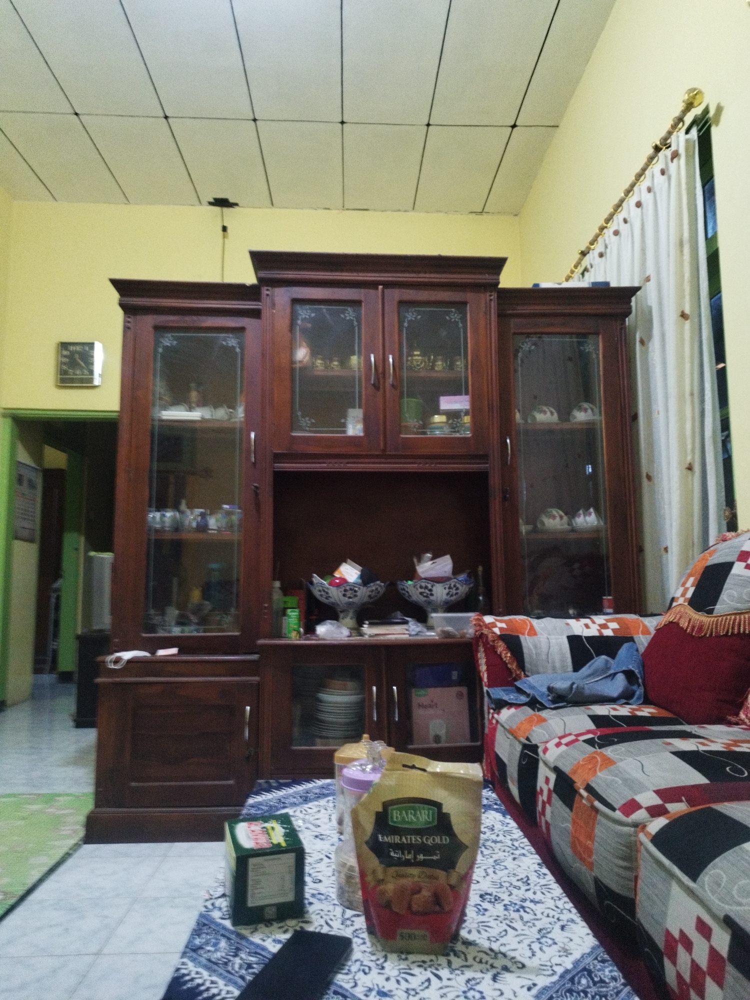 The image depicts an indoor scene, likely a living room, with a large wooden cabinet dominating the center. The cabinet has multiple glass-fronted sections displaying various items, including what appear to be decorative plates and glassware. To the right of the cabinet is a sofa with a colorful, patchwork-style upholstery. In the foreground, on a patterned rug, is a bag of "BARARI Emirates Gold" dates and a green box. A partially visible doorway with a green frame is visible to the left. The walls are painted yellow, and the ceiling is a grid of white tiles with visible black lines. A wall clock is mounted on the left wall. The overall impression is of a furnished domestic interior. The image does not provide any specific clues to pinpoint a location within Sumenep, Indonesia, beyond it being an indoor domestic setting.