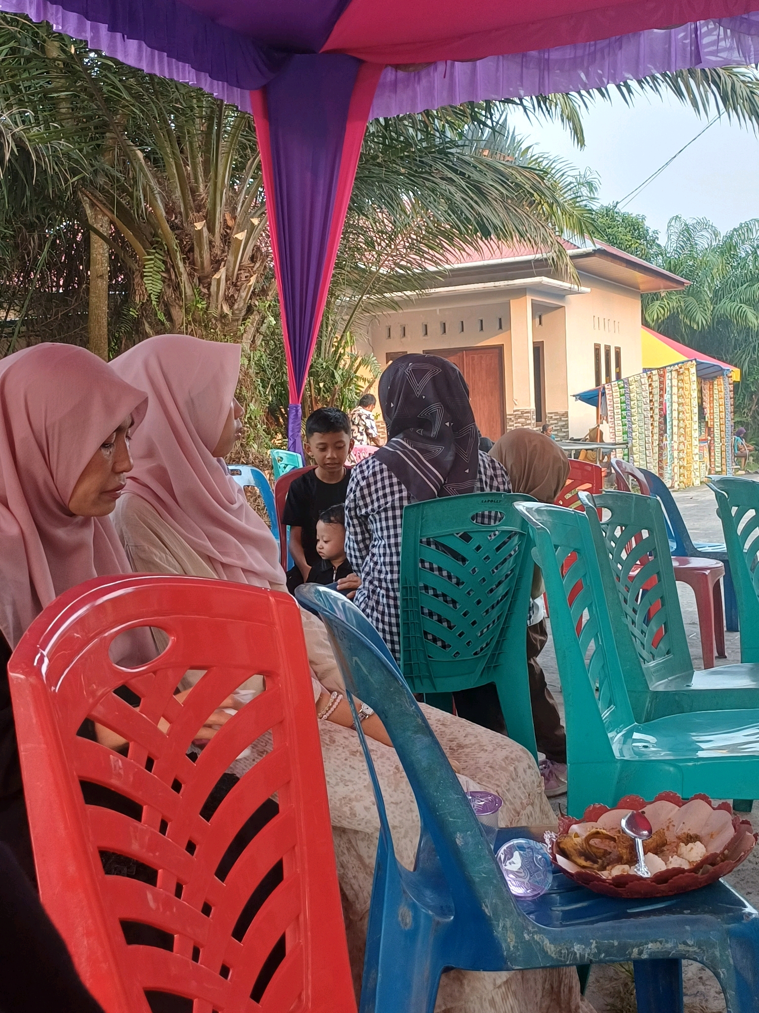 An outdoor gathering is taking place under a purple and pink tent. In the foreground, a red plastic chair is visible, with a blue plastic chair to its right. On the blue chair, a red decorative bowl contains food, possibly fried items and rice, with a small spoon. To the left, two women wearing pink hijabs are seated, one looking forward and the other with her head bowed. Behind them, a young boy in a black t-shirt sits, holding a younger child. Further back, a person wearing a black and white checkered headscarf and outfit is seated. In the background, a simple house with a red roof is visible, surrounded by palm trees and other vegetation. The ground appears to be paved or concrete. A row of colorful items, possibly snacks or small goods for sale, is displayed to the right of the house. The overall scene suggests a community event or celebration in a residential area of Dumai City, Indonesia.