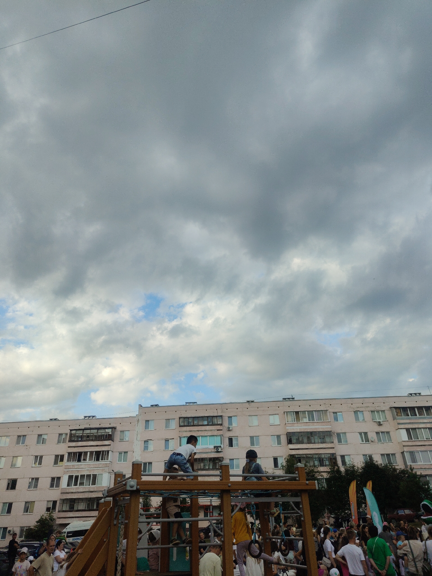 The image depicts a cloudy sky over a residential building in Mendeleyevsk, Russia. In the foreground, a playground structure is visible with children on it. One child, wearing a white t-shirt and blue shorts, is climbing on the monkey bars. Another child, in a white t-shirt and dark shorts, is also on the structure. A crowd of people is gathered in the background, some observing the children on the playground. The residential building behind the playground is a multi-story apartment complex with many windows. Trees and some vehicles are also visible.