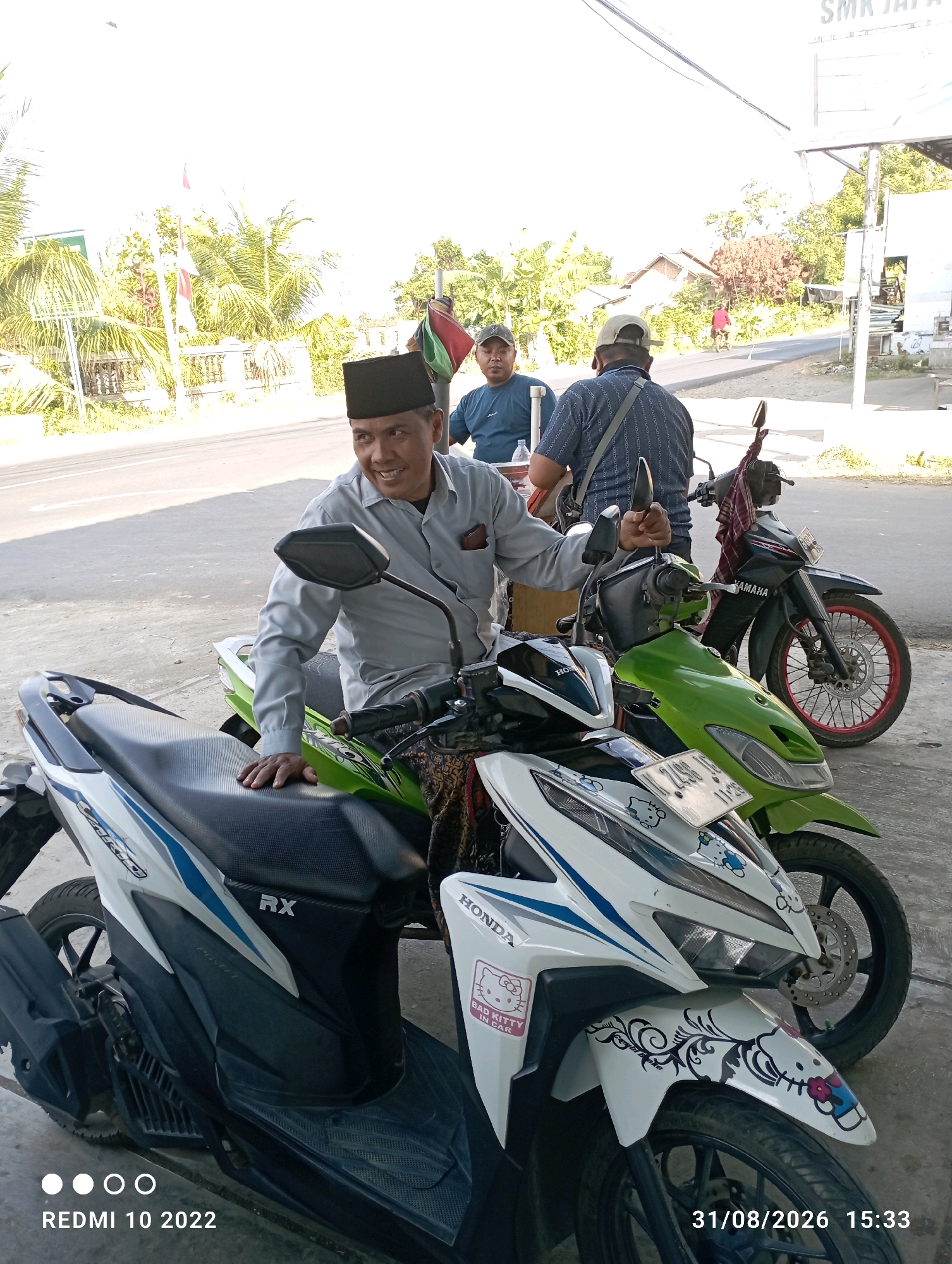 In Dukuhseti, Indonesia, three men are gathered near several parked motorcycles under a bright sky. The central figure, wearing a light grey collared shirt and a black peci (a cap), is seated on a white and blue scooter, looking to his left and smiling while holding a small object in his right hand. To his left, a man in a navy blue t-shirt and a cap is partially visible, seated and looking towards the center. Behind the central figure, another man in a dark patterned shirt and a beige cap is standing. Two green motorcycles and a black Yamaha motorcycle are parked alongside the scooters. In the background, a road is visible with trees, buildings, and a white sign with text that appears to say "SMK JAYA". The overall environment suggests an outdoor gathering or a stop along a road. The image also contains timestamps "REDMI 10 2022" and "31/08/2026 15:33".