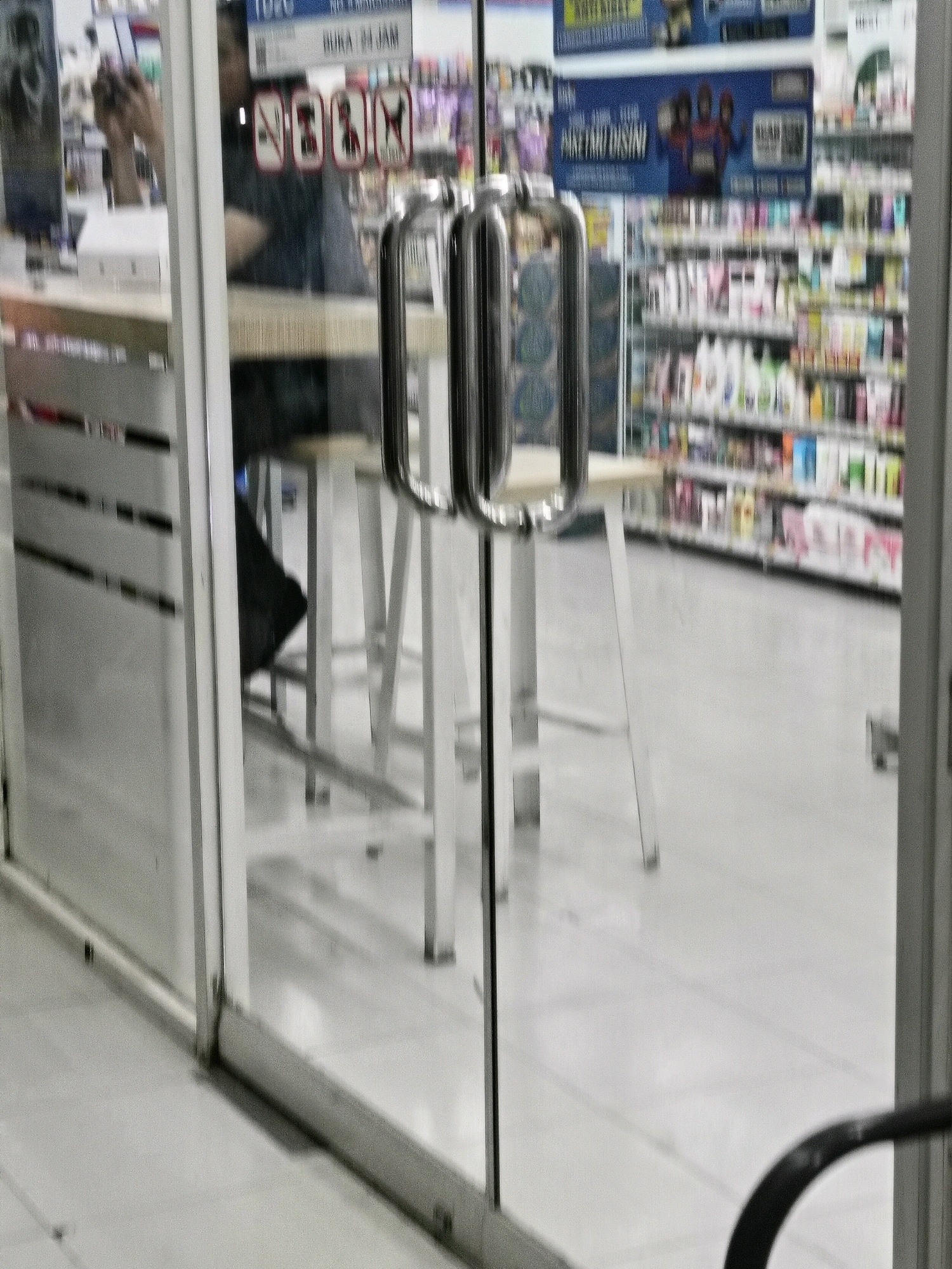 The image shows the interior of a brightly lit convenience store, likely a Circle K or similar establishment, as suggested by the branding and store layout visible through the glass doors. The doors are partially open, revealing a person dressed in dark clothing reflected in the glass, holding a phone to their ear. The interior features shelves stocked with various products, including beverages and personal care items, common in Indonesian convenience stores. The floor is tiled and appears clean. The overall impression is a typical retail environment in an urban setting. The location is identified as Makassar, Indonesia.