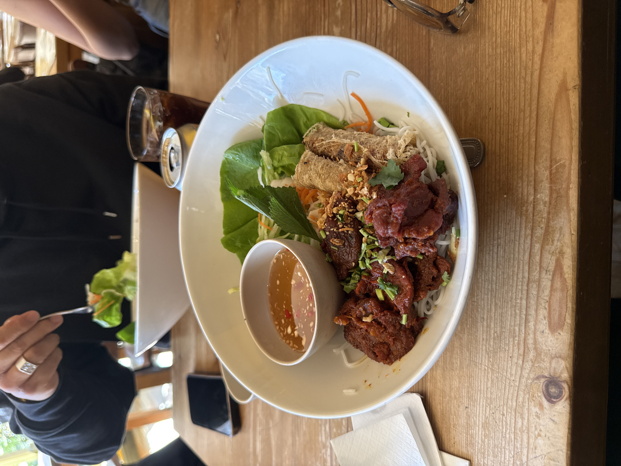 A top-down, eye-level shot features a partially eaten dish of Vietnamese vermicelli salad, also known as Bun Cha, on a light wood table. The dish is in a white bowl and contains vermicelli noodles, sliced lettuce, spring rolls, grilled pork, and a small bowl of dipping sauce. A can and a glass of what looks like cola are visible to the left of the bowl. A pair of eyeglasses rests on the table's top-right corner. A man holding a fork is visible on the left edge, lifting some salad. There are white napkins on the lower part of the frame. The scene suggests an informal dining setting. The location cannot be determined.