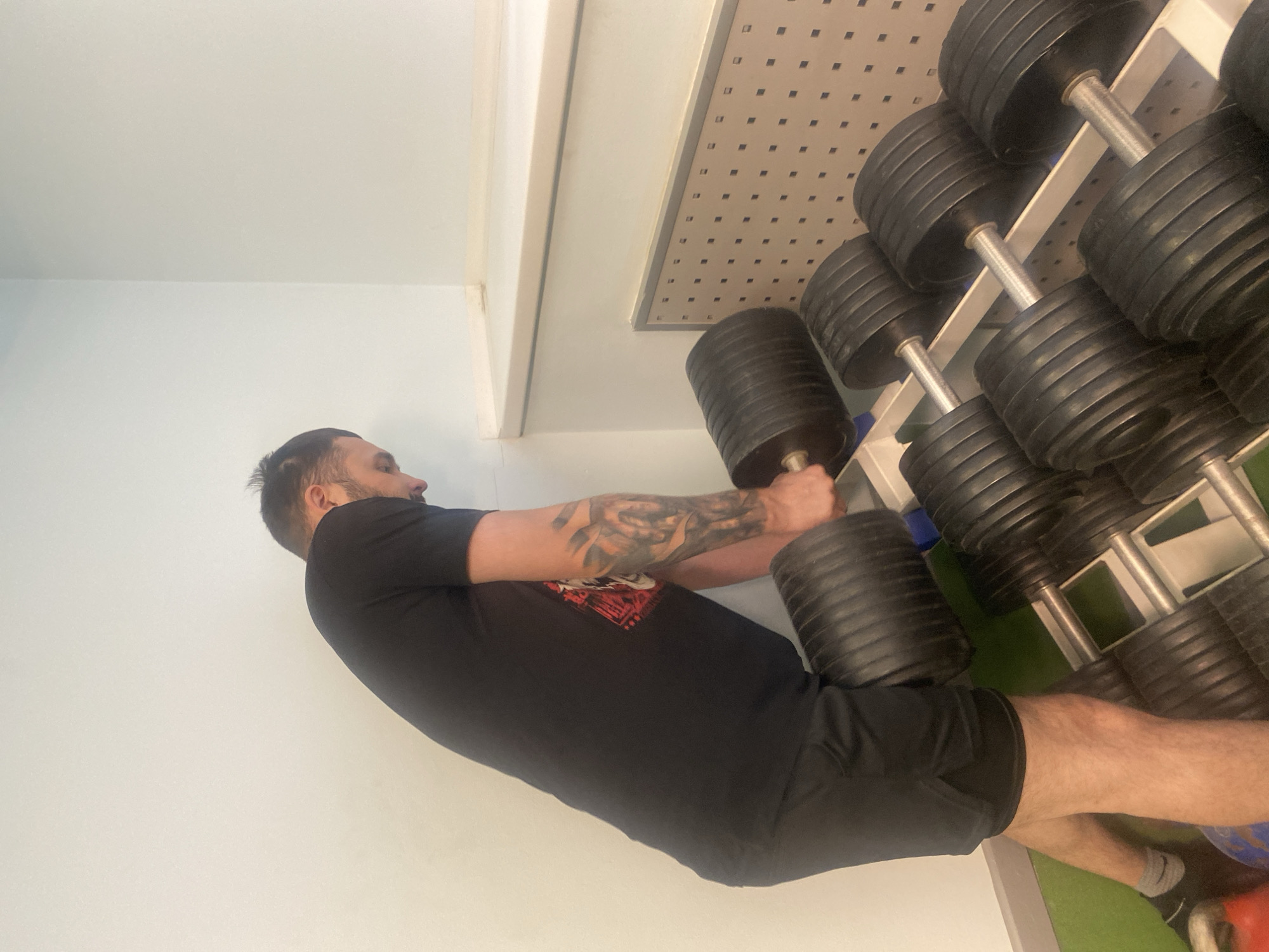 A medium shot shows a man in a gym in Chaikovsky, Russia. The man is light-skinned with short brown hair, a beard, and a tattoo on his left forearm. He is wearing a black t-shirt and black shorts. He is standing in front of a rack of dumbbells, and he is holding a large dumbbell in both hands, ready to lift it. The dumbbells are black and silver. The background is a light blue wall and a white ceiling with a perforated panel. There is a green floor. The scene is well-lit.
