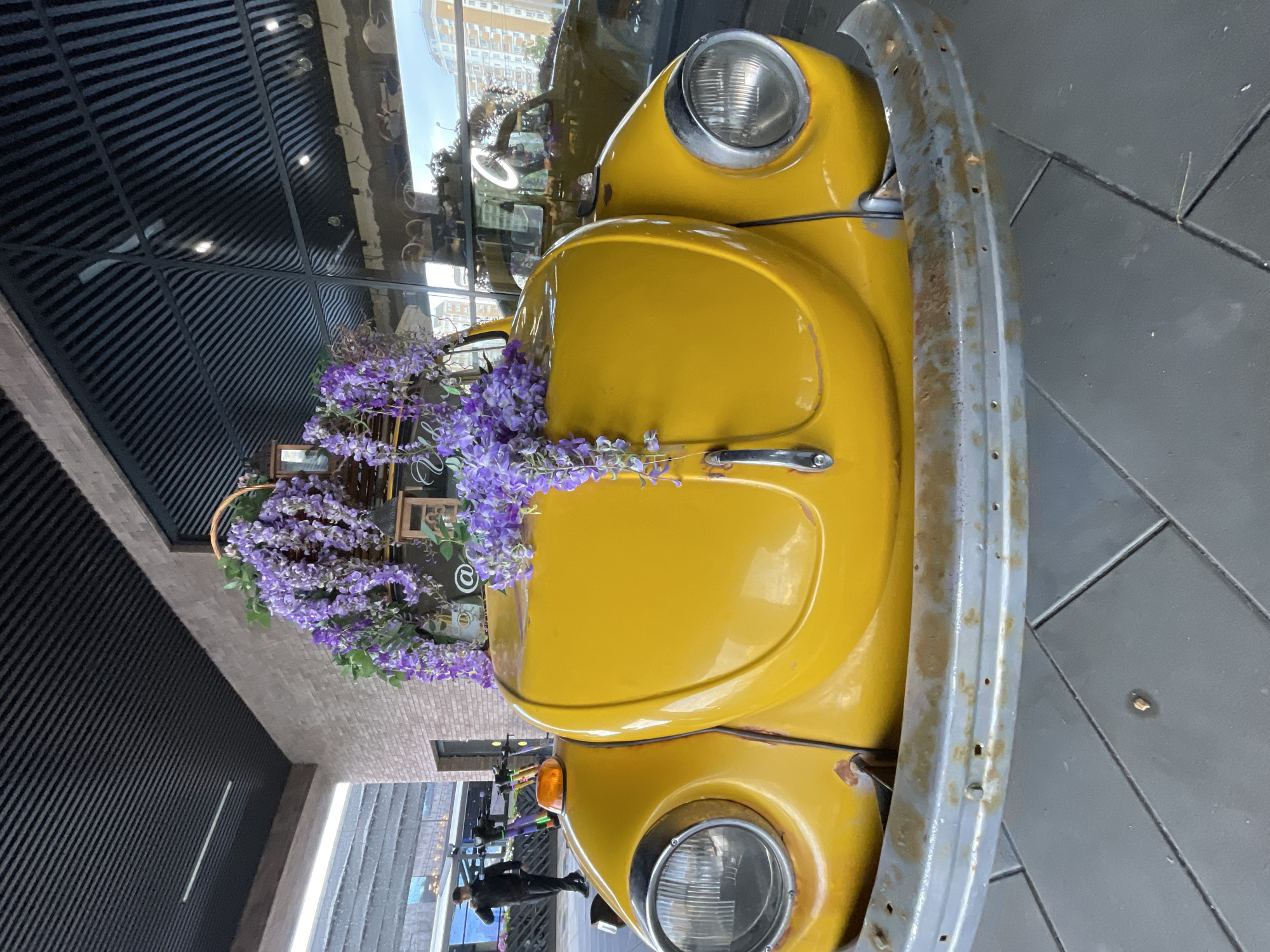 A yellow Volkswagen Beetle, decorated with cascading purple flowers, is parked outdoors. The car has a rusty chrome bumper and a prominent headlight. In the background, a person in dark clothing is walking, and a building with a brick facade is visible. The scene appears to be in a modern urban environment, possibly a street or plaza.