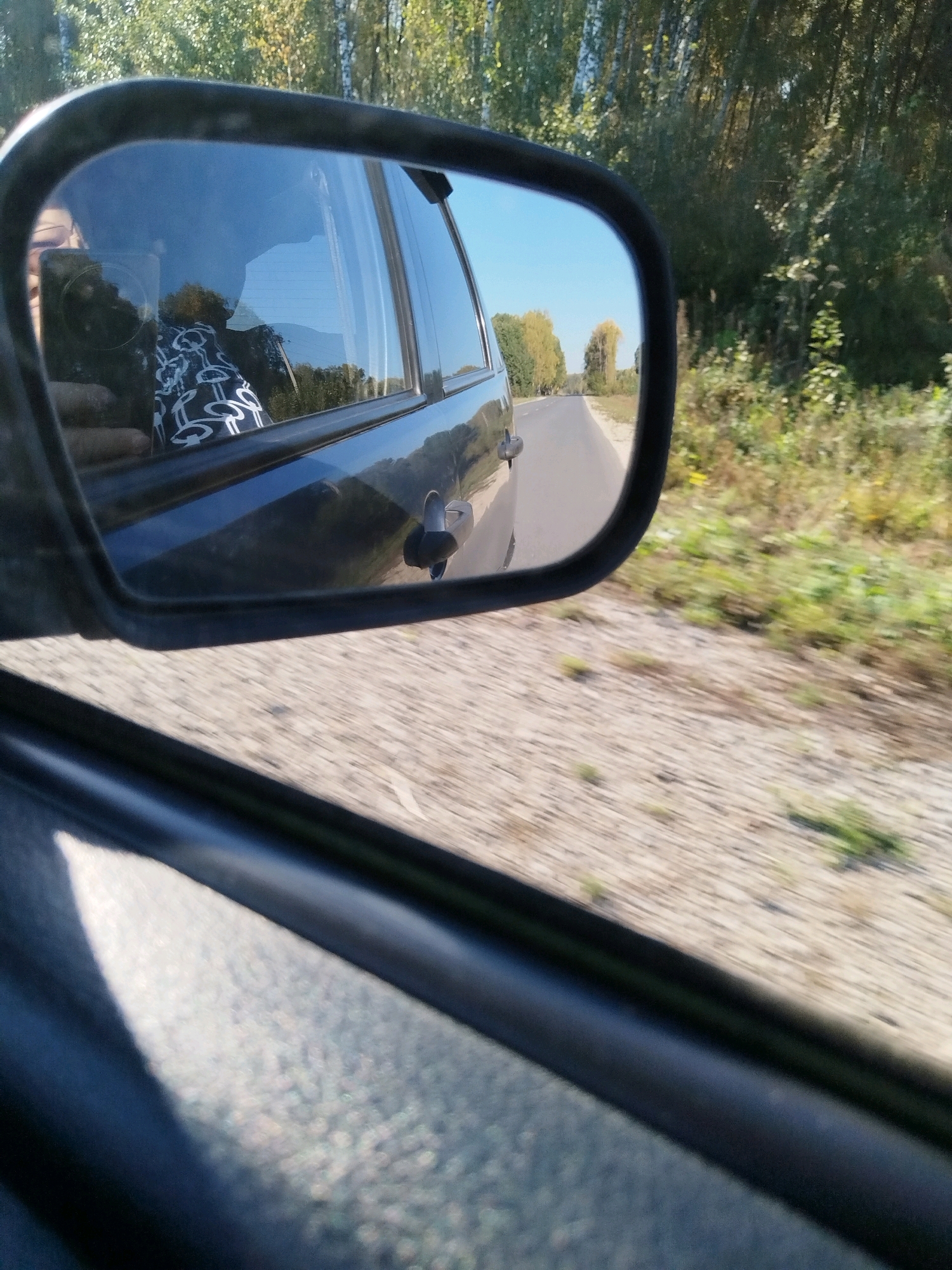 The image shows a reflection in a car's side mirror, capturing a person holding a smartphone, a person's arm, and the interior of the car, likely a dark-colored sedan. The mirror also reflects a road stretching into the distance, bordered by trees and vegetation. The environment appears to be rural, with a clear blue sky overhead. The overall impression is of a journey on a sunny day, possibly in the vicinity of Novo-Kuskovo, Russia, given the context. The image is taken from inside the car, looking out through the side window towards the mirror.