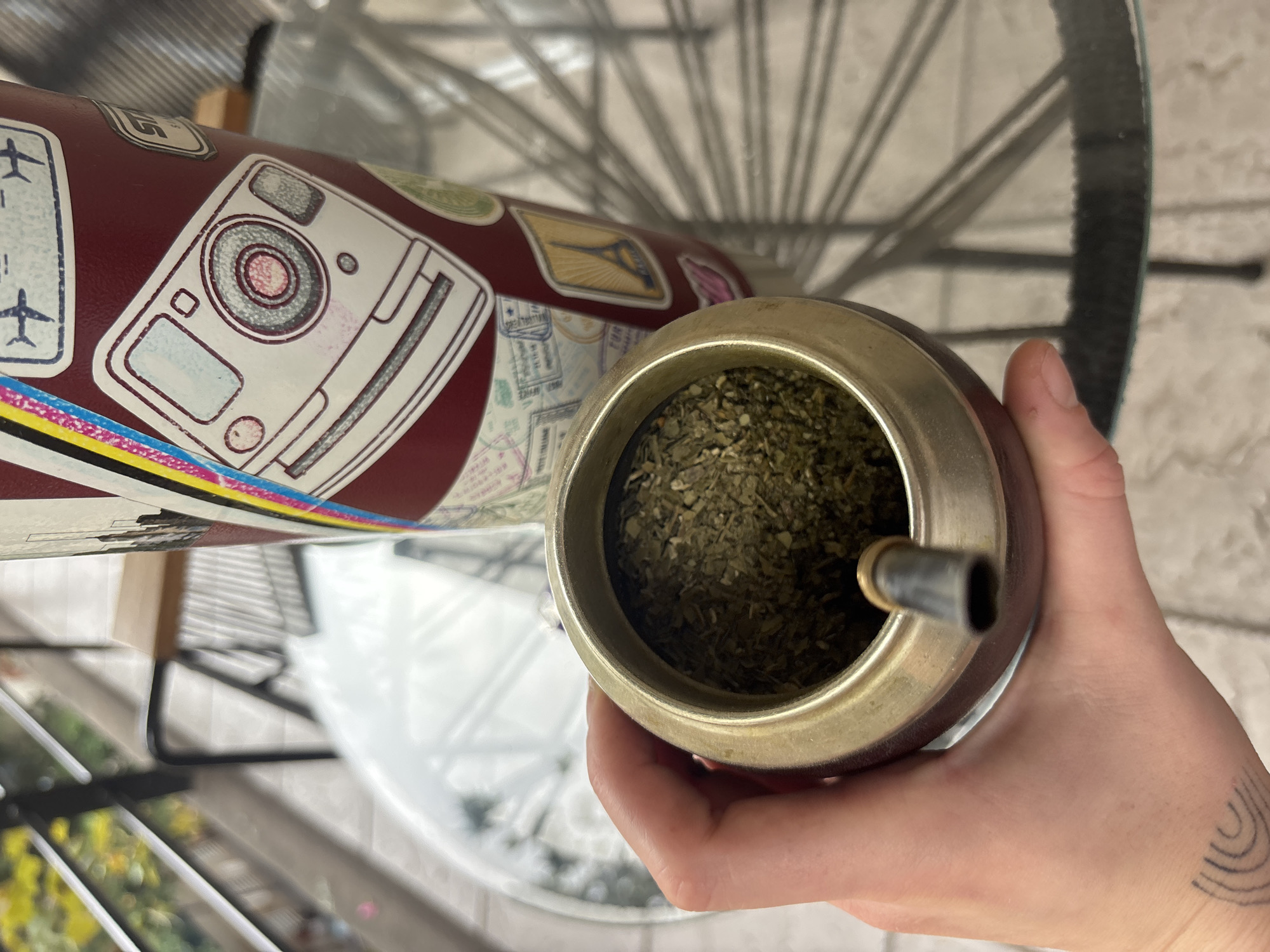 The image captures a close-up, high-angle view of a person's hand holding a traditional mate gourd, positioned next to a travel-themed thermos, likely in Buenos Aires, Argentina.
The central subject is a dark red or maroon mate gourd with a metal rim, filled with green, finely chopped yerba mate. A metallic bombilla (straw) is inserted into the mate, indicating preparation for drinking. A light-skinned hand, with an abstract tattoo of concentric arcs on its outer side, gently holds the gourd.
Beside the gourd stands a large, dark red thermos (termo) covered in an array of travel-related stickers. Notable stickers include a white, stylized vintage camera with red and grey accents, an airplane, and various passport stamps. Some stamps feature illegible text, while one clearly shows "EUROPE" and partially visible numbers like "S201440." Another sticker depicts a landmark resembling the Eiffel Tower. The bottom of the thermos is accented with a colorful band of blue, yellow, and pink stripes.
The background is softly blurred, revealing a glass table surface. Reflected and visible through the glass is the intricate, spoked structure of a metallic object, possibly a chair or part of a railing, casting faint shadows. The lighting suggests it is daytime. The image conveys a sense of a common, relaxed daily ritual.
