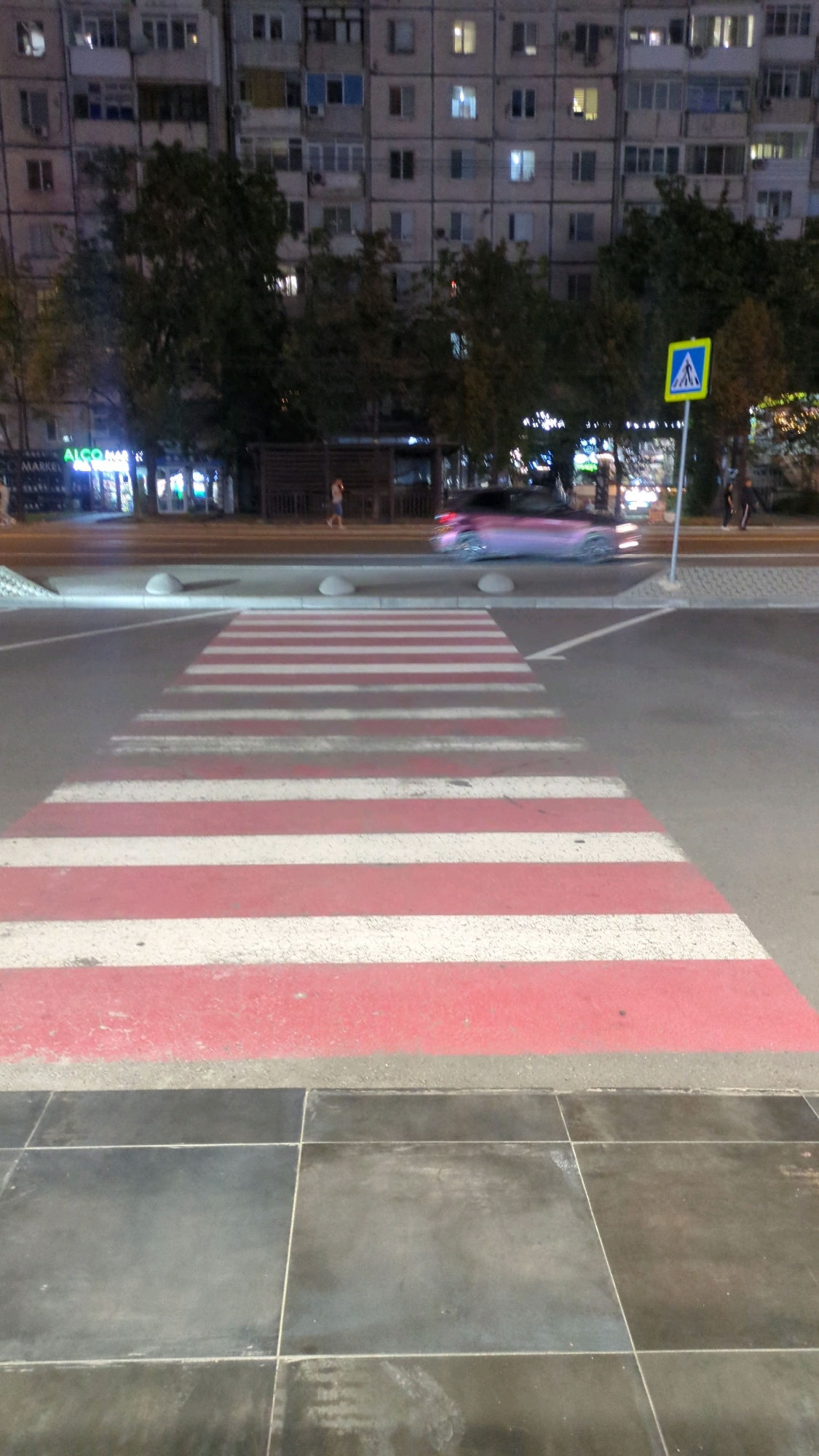 This is a nighttime photograph of a pedestrian crossing on a city street in Chișinău, Moldova. The pedestrian crossing is marked with alternating red and white stripes and is the central focus of the foreground. To the left of the crossing, there is dark tiled pavement. In the background, a multi-story apartment building with illuminated windows is visible. Several trees line the street between the building and the road. A crosswalk sign with a blue background and a white pedestrian symbol is positioned on the right side of the image, near the road. A car, blurred due to motion, is driving across the pedestrian crossing. Two people are standing on the sidewalk to the right of the sign, and another person is walking on the sidewalk further to the left, near the buildings. The scene appears to be a typical urban street environment at night.